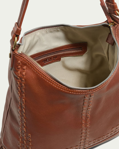 The Evelyn Hobo by American Leather Co. is a brown leather handbag with visible stitching along the sides. It opens to an off-white interior lining and includes a zippered pocket trimmed in brown leather. Made from genuine leather, this handbag features a shoulder strap with brass hardware for a chic touch.