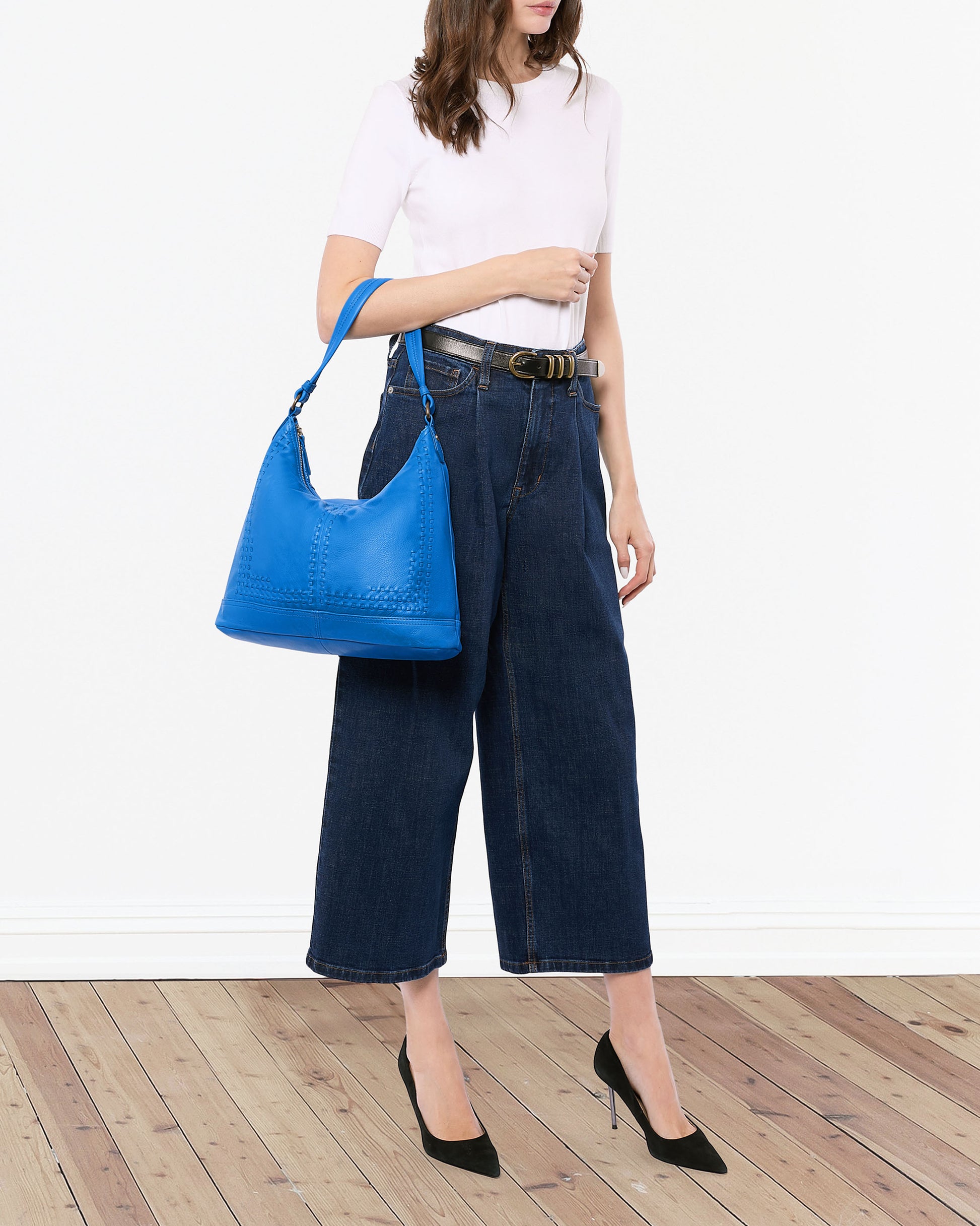 A woman in a white top and wide-leg jeans stands on a wooden floor, showcasing an Evelyn Hobo from American Leather Co. She sports black heels, her hair cascading down, emphasizing the luxury of the hobo bag against a plain white background.