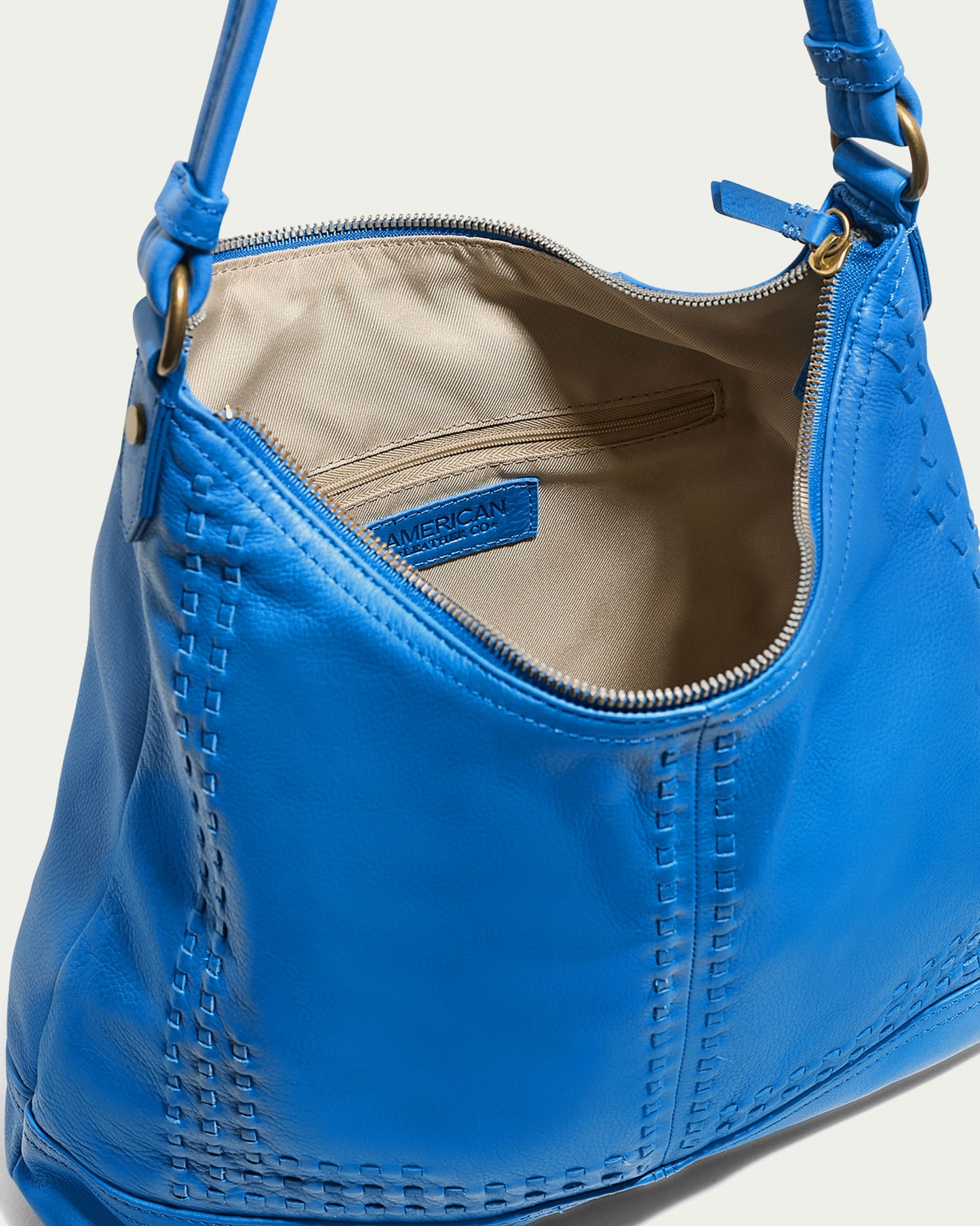 The Evelyn Hobo from American Leather Co. is an exquisite hobo bag made from premium American leather in a striking blue color. It features intricate decorative stitching and a woven texture, complemented by a zippered opening that leads to a beige interior lining with an internal zip pocket. A single strap, elegantly fastened with brass rings, completes the design.