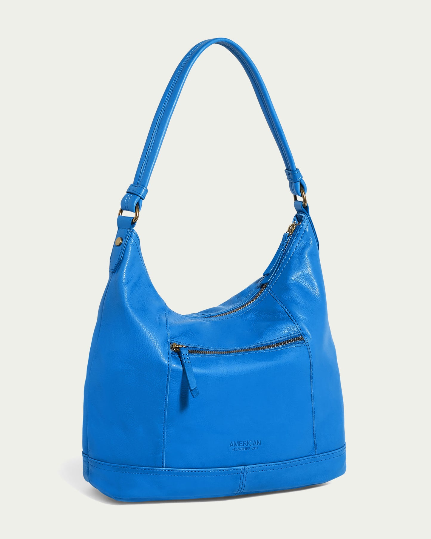 Introducing the Evelyn Hobo by American Leather Co., a luxurious handbag made from rich American leather. This bright blue, single-strap bag showcases a front zipper pocket and subtle stitching details. Its smooth texture and small embossed brand logo add to its elegance against an off-white backdrop.