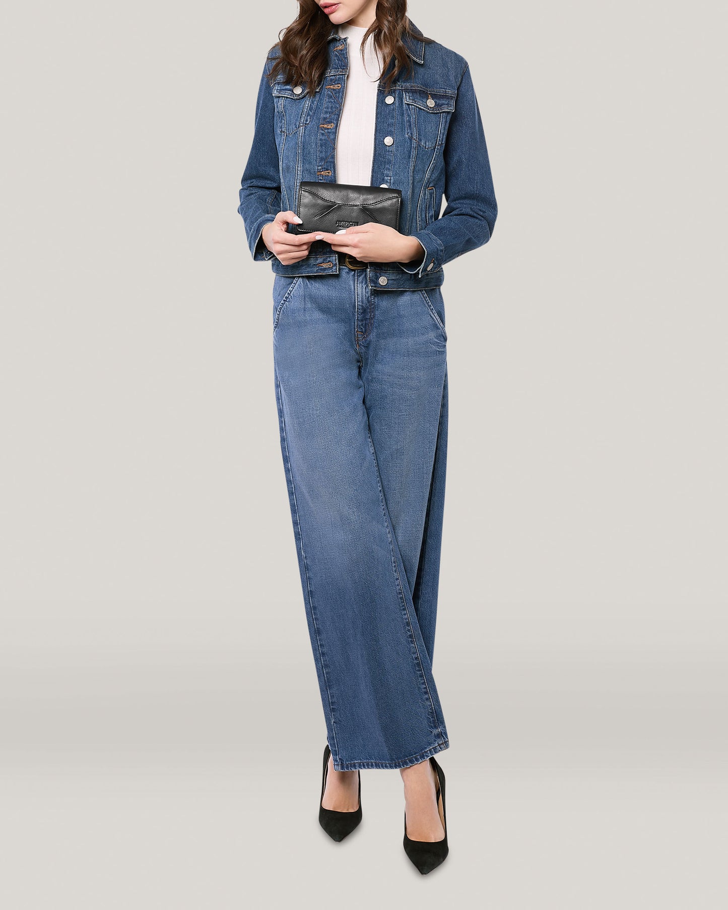 A person wearing a blue denim jacket, white shirt, and wide-leg blue jeans holds an American Leather Co. Hickory Bifold Wallet in front of a plain light gray background. They are standing with legs crossed and wearing black high-heeled shoes. The person's face is not visible.