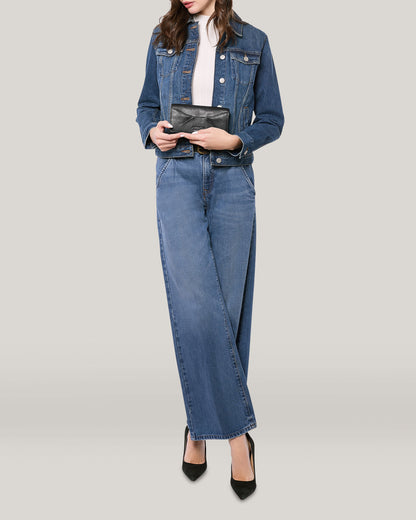 A person wearing a blue denim jacket, white shirt, and wide-leg blue jeans holds an American Leather Co. Hickory Bifold Wallet in front of a plain light gray background. They are standing with legs crossed and wearing black high-heeled shoes. The person's face is not visible.