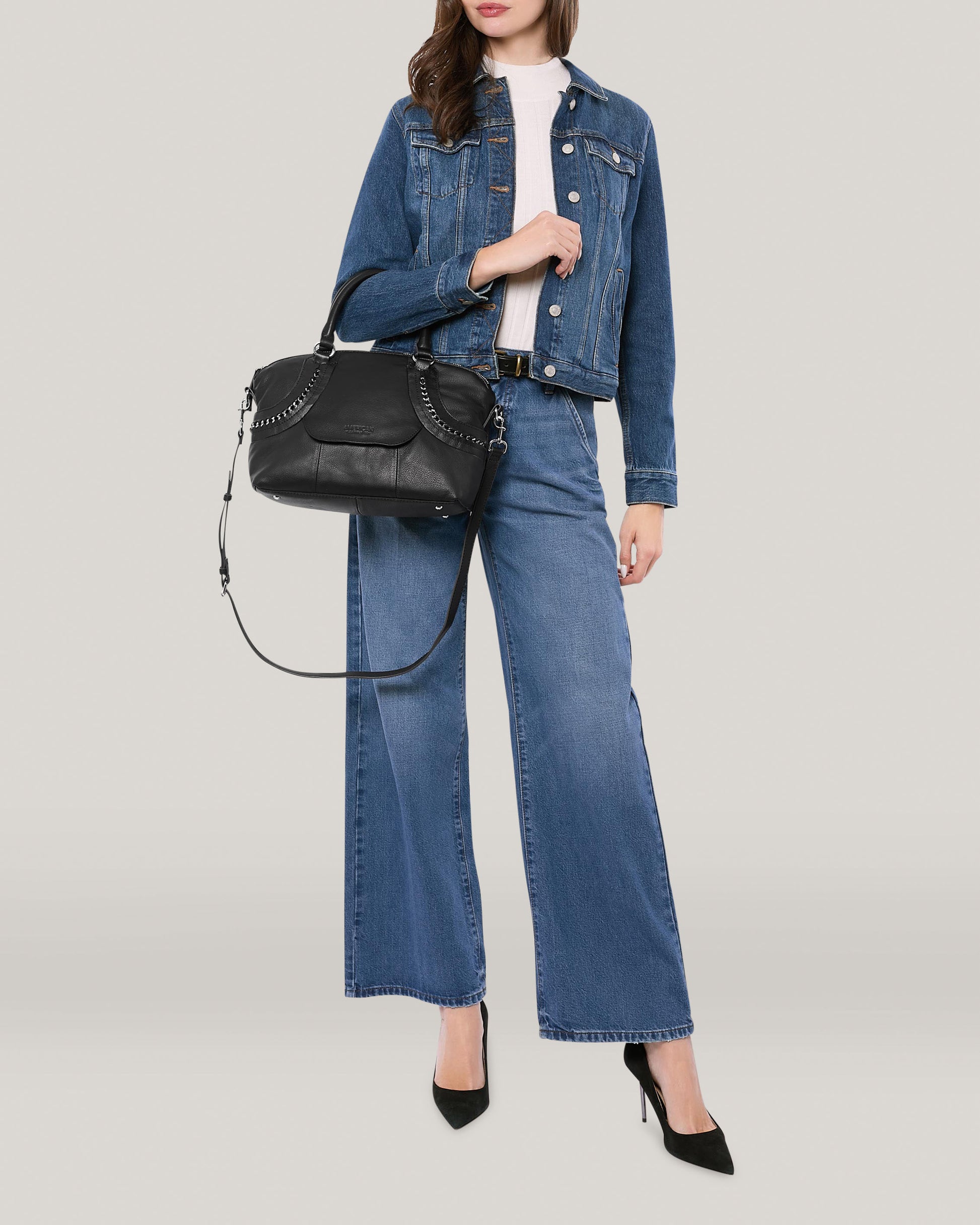 A person wearing a denim jacket and wide-legged jeans poses against a light background. They hold the Janssen Dome Satchel by American Leather Co., crafted from genuine leather, and wear black high heels. Only the lower half of their face is visible.