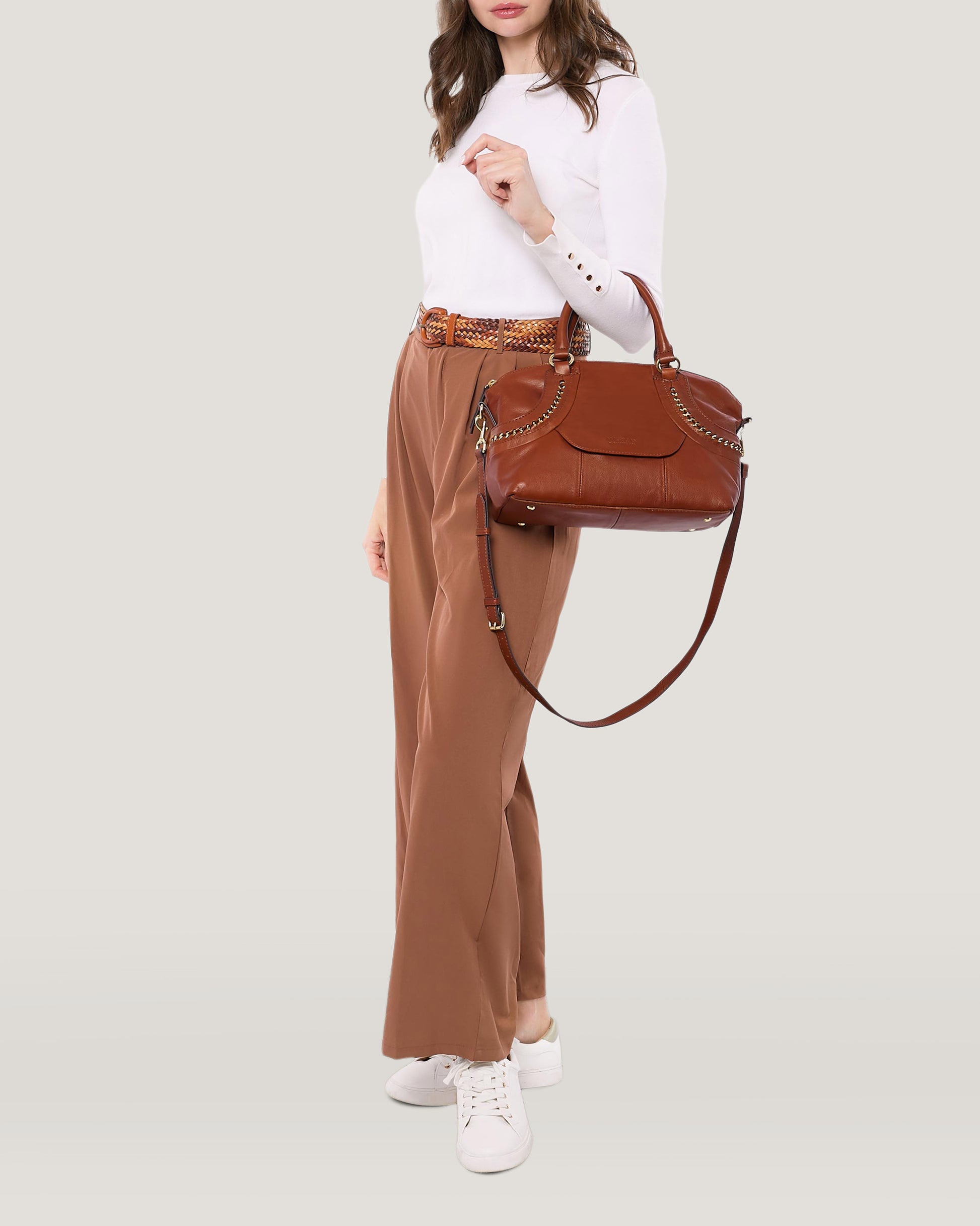 A person is dressed in a white long-sleeve top and brown trousers, carrying an American Leather Co. Janssen Dome Satchel, which features a shoulder strap. They are also wearing white sneakers and have their trousers secured with a patterned belt against a plain light gray background.