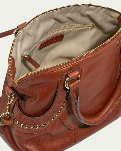 The Janssen Dome Satchel by American Leather Co. is a genuine leather handbag in brown, embellished with gold chain accents. It features a zippered top that opens to a beige interior, complete with a small pocket and label. This elegant bag offers versatile carrying options with both handles and a detachable shoulder strap.