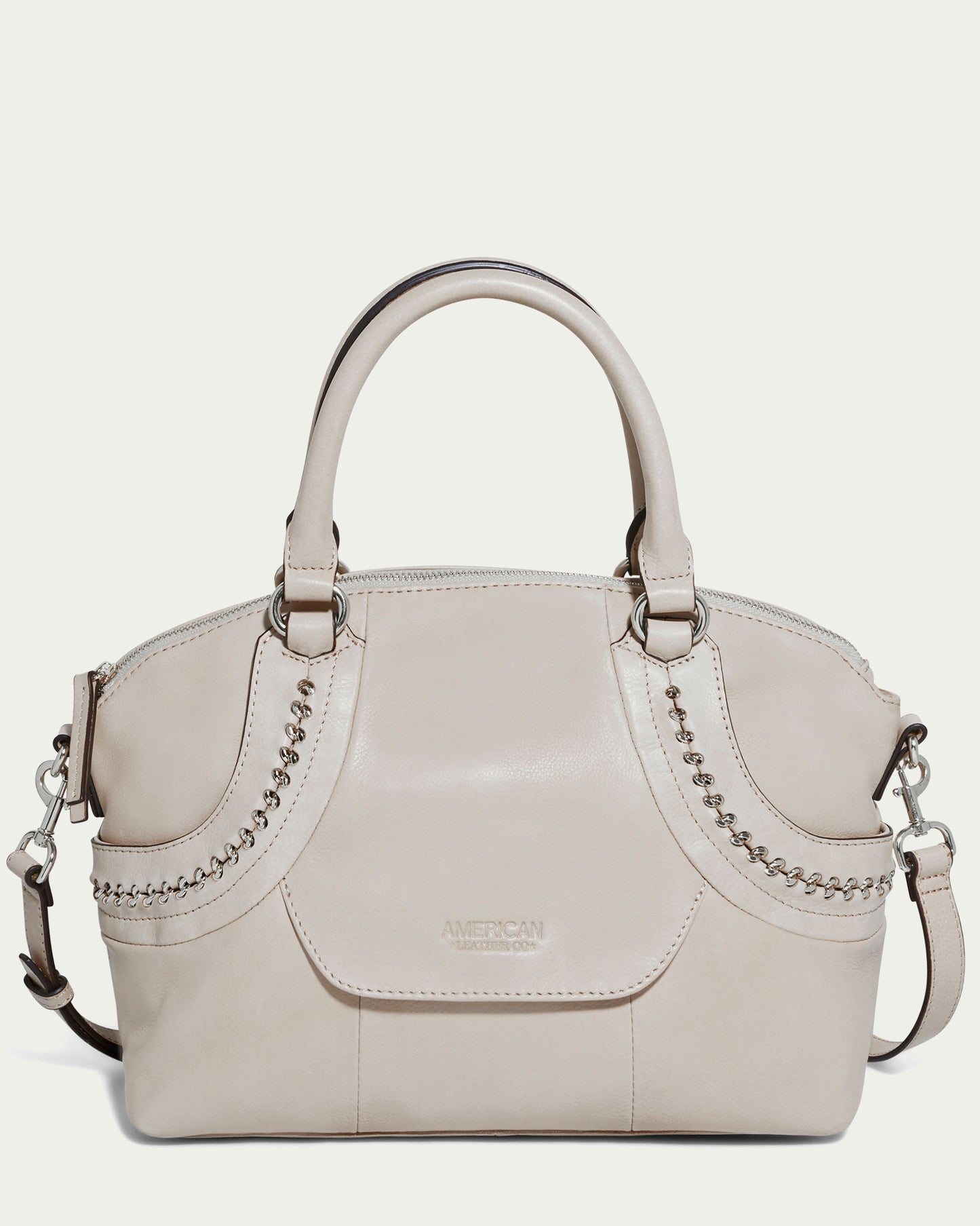 The Janssen Dome Satchel from American Leather Co. is a cream-colored leather bag that showcases decorative stitching and metal chain accents. It includes two handles, an adjustable shoulder strap, and a small front pocket embossed with "American," combining elegance with practicality in its design.