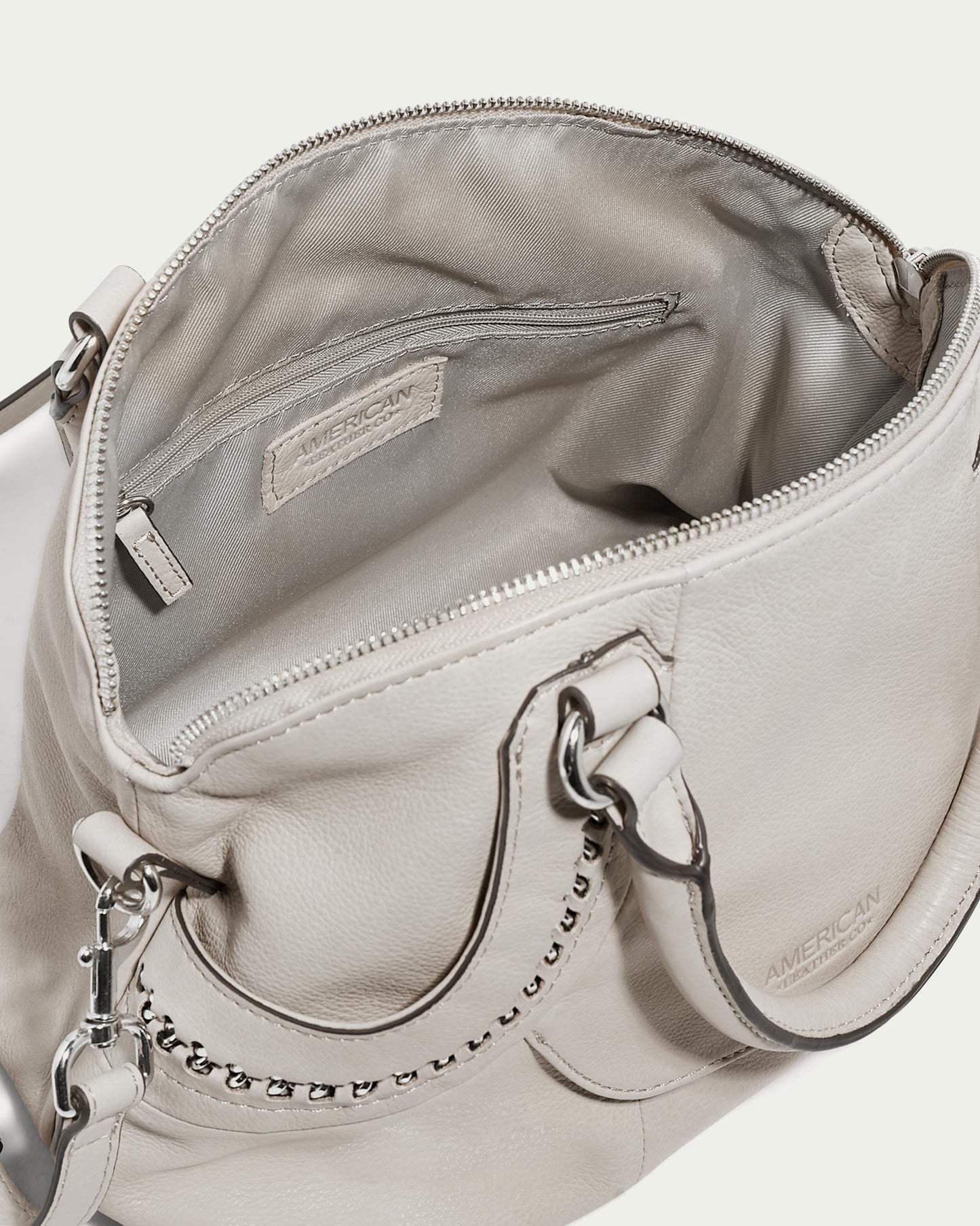 The Janssen Dome Satchel by American Leather Co. is a light grey leather bag with a zippered top that reveals its spacious interior. It features metal chain and leather handles, showcasing its high-quality construction. Inside, an inner fabric tag can be seen, and it includes an adjustable shoulder strap for added versatility.