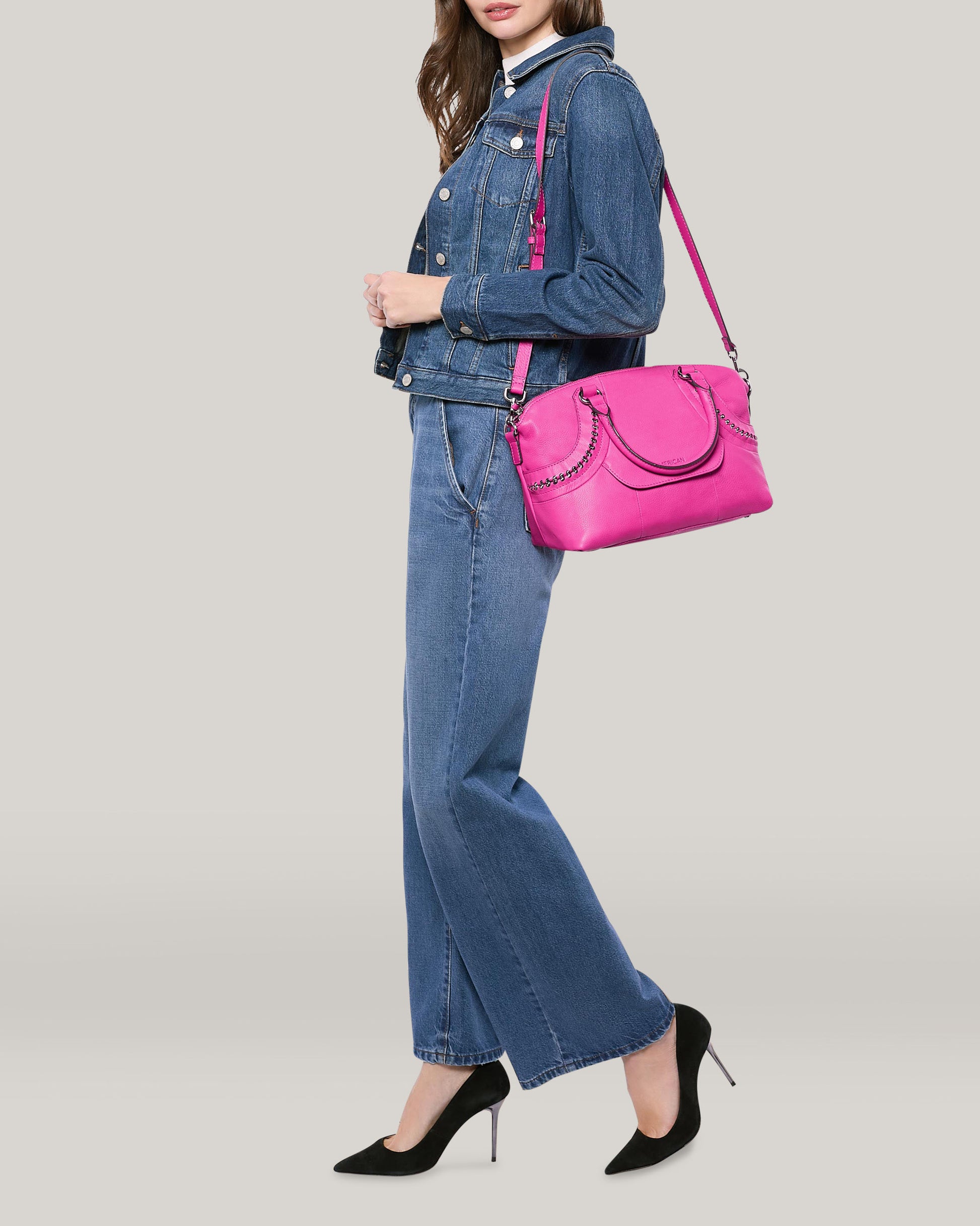 A person dressed in a denim jacket and jeans is carrying an American Leather Co. Janssen Dome Satchel in pink on their shoulder. They are set against a plain background, wearing black heels.