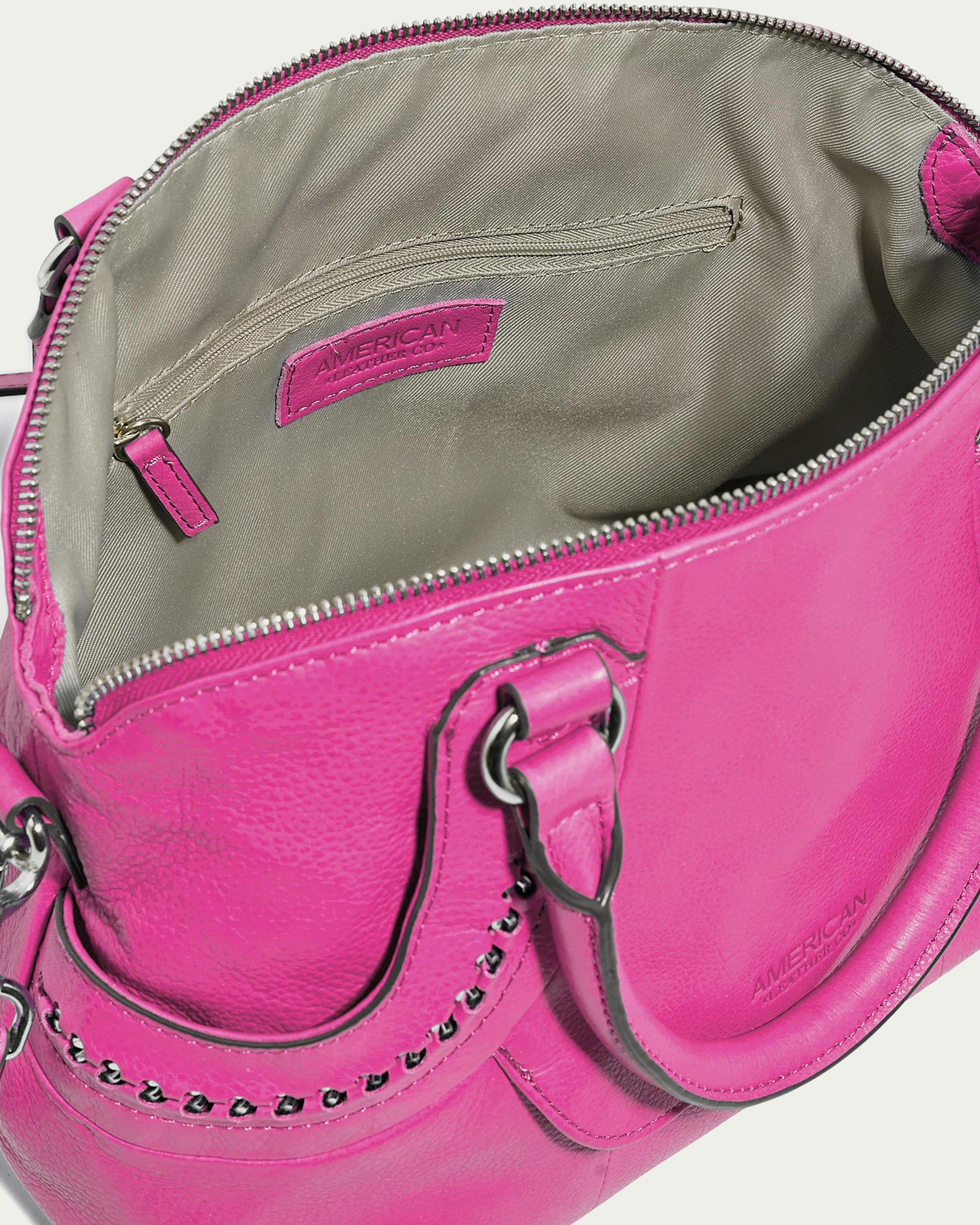 A top-down view of the pink Janssen Dome Satchel featuring silver hardware displays the open bag with its beige inner lining and zippered pocket. The brand name "American Leather Co." is embossed inside, making it an ideal everyday leather bag.