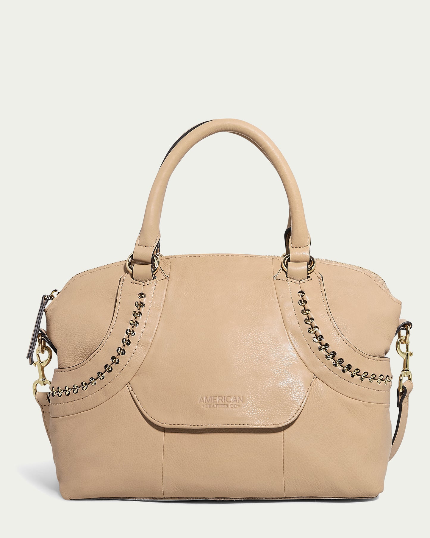 The Janssen Dome Satchel by American Leather Co. is a chic curved handbag crafted from beige genuine leather. It features two short handles, decorative chain stitching, gold-tone hardware, and a branded logo on the front, making it a stylish choice for everyday use.