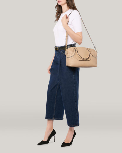 A person wearing a white t-shirt and blue high-waisted jeans poses with the Janssen Dome Satchel from American Leather Co. elegantly draped over their shoulder. The beige genuine leather handbag complements their black high heels as they stand against a plain light gray background.