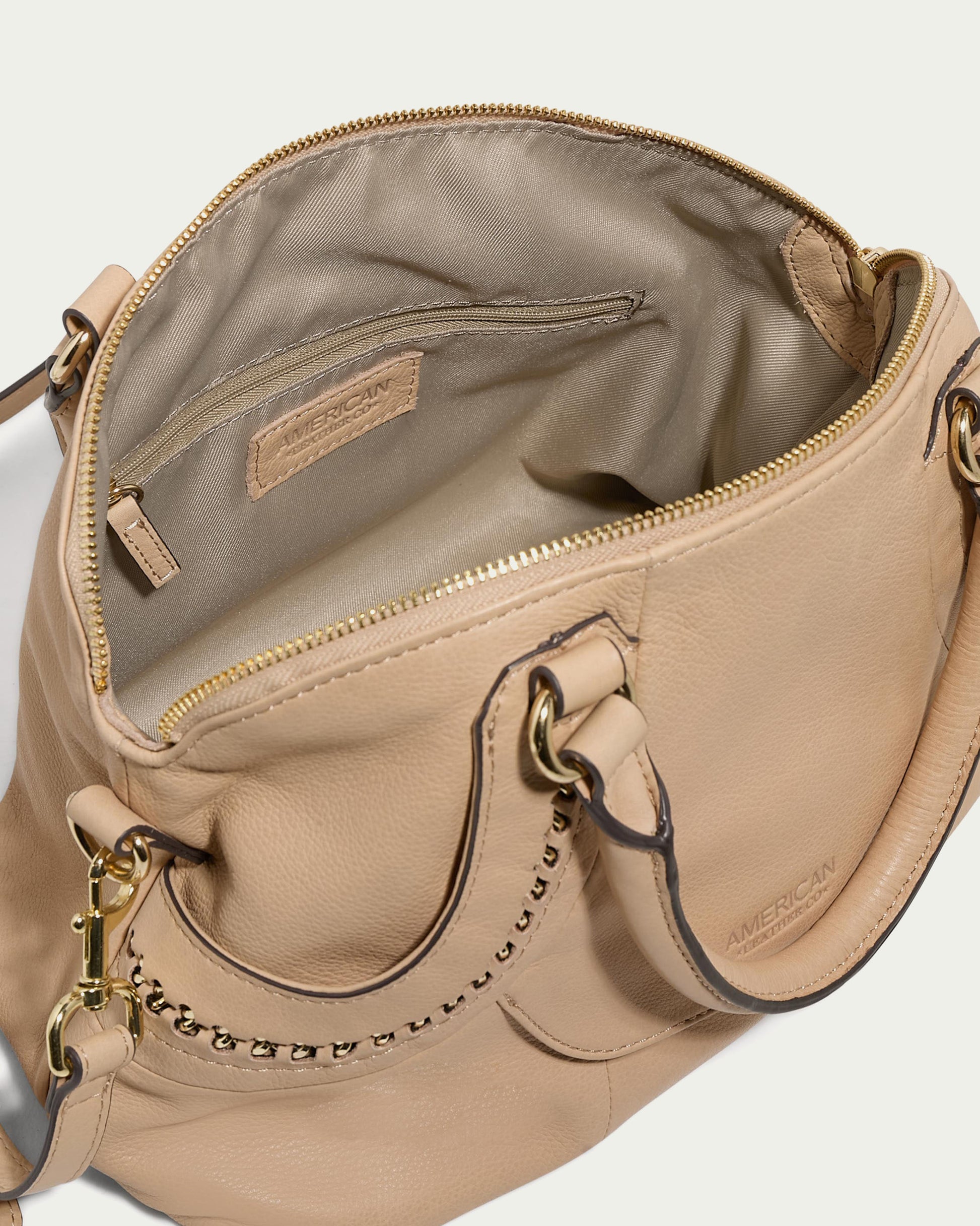 The Janssen Dome Satchel by American Leather Co. is a beige leather handbag featuring a zippered top that opens to reveal a roomy interior. Made from genuine leather, it includes a detachable strap with gold hardware and decorative chain accents. Inside, the brand name is embossed on a leather patch, making it an ideal everyday bag.