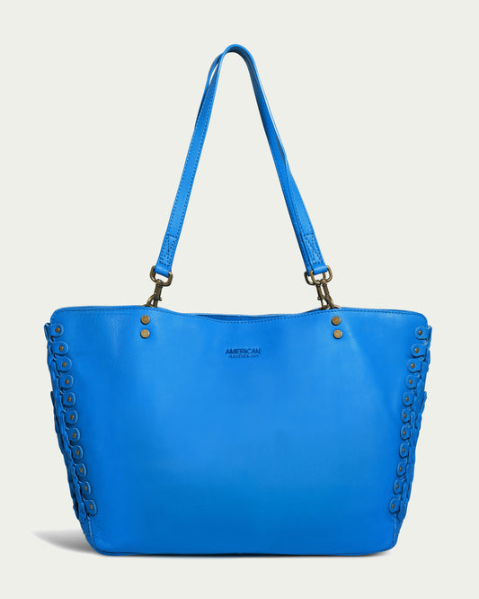 The Lenox Triple Compartment Tote by American Leather Co. is a cerulean blue leather tote bag with double shoulder straps and metallic rivet detailing along the sides. Made from genuine leather, the bag has a simple design, featuring a small logo stamped on the front center against a plain, light-colored background.