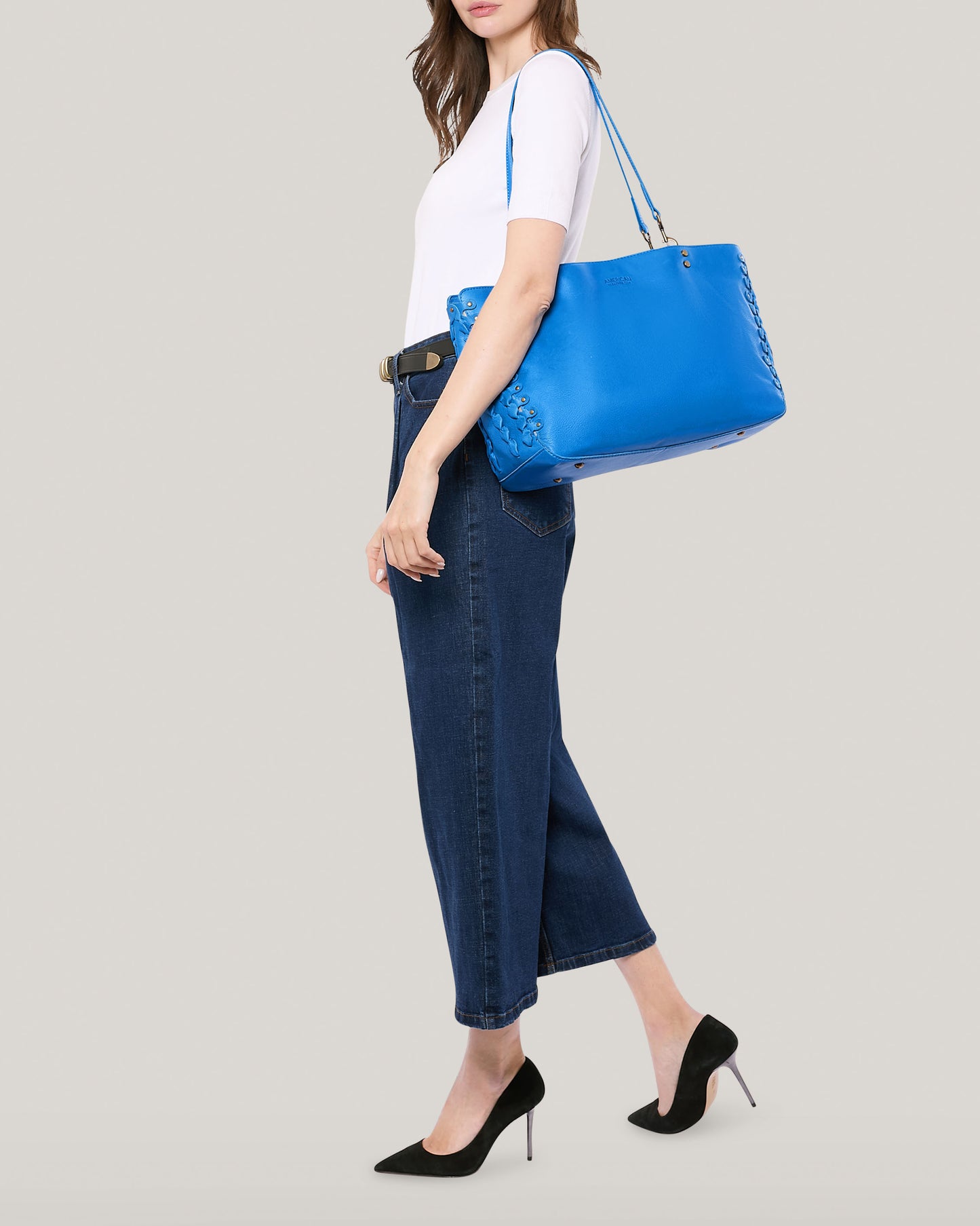 A woman stands against a neutral background, carrying the Lenox Triple Compartment Tote by American Leather Co. on her shoulder. She is wearing a white t-shirt, blue high-waisted jeans, and black high-heeled shoes. Her head is slightly turned to the side, and her right arm hangs by her side.