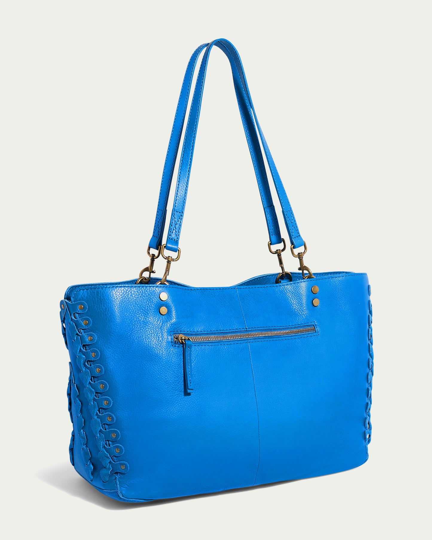 The Lenox Triple Compartment Tote by American Leather Co. is a large, blue genuine leather bag with double shoulder straps and brass hardware. It features a zippered pocket on the front, decorative lacing along the sides, and a triple entry compartment for added convenience. The background is plain off-white.