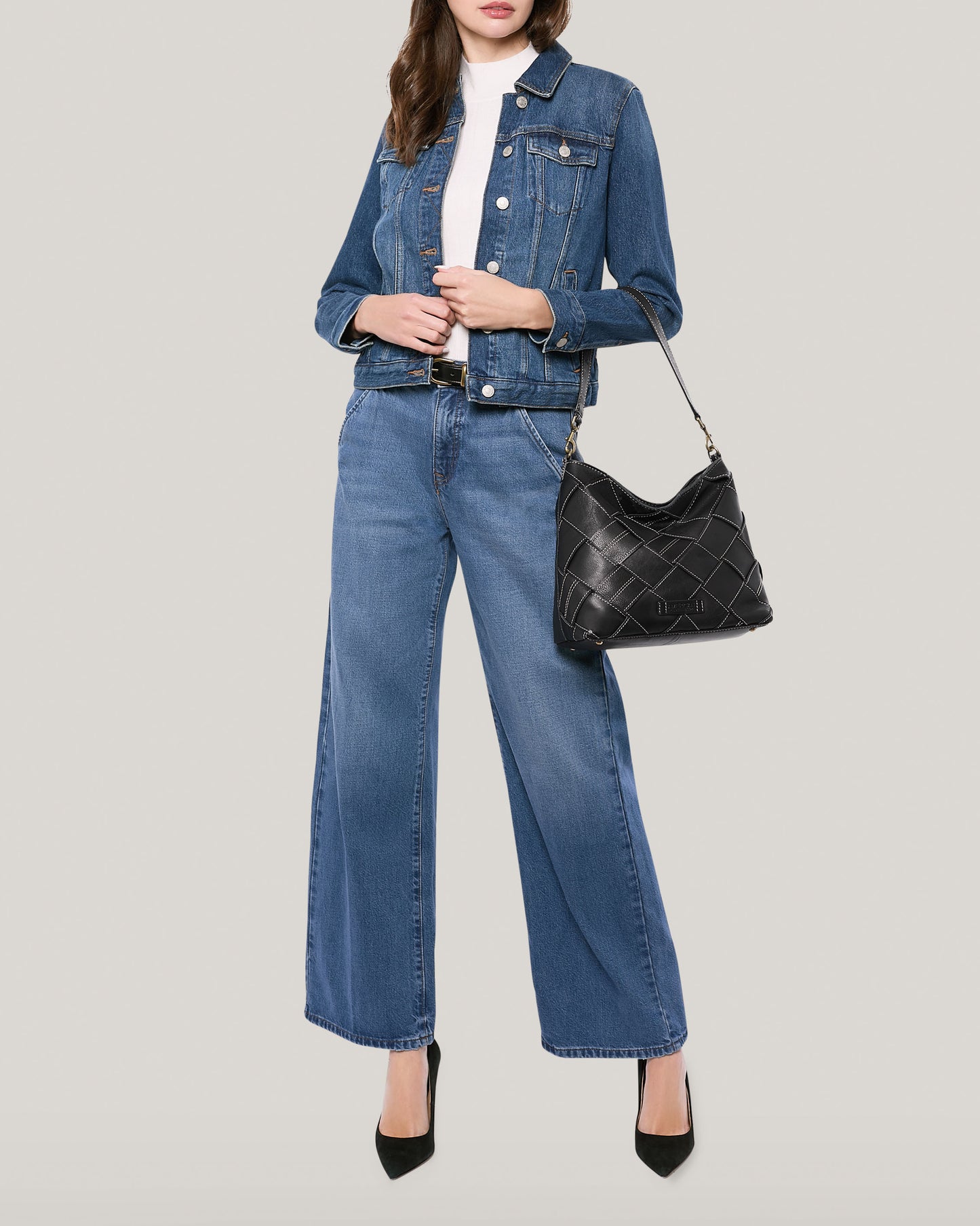 A woman dressed in a blue denim jacket and wide-leg jeans poses against a light backdrop, carrying an American Leather Co. Mercer Shoulder handbag with a detachable shoulder strap on her shoulder while wearing black high heels. Her hands rest in her pockets.