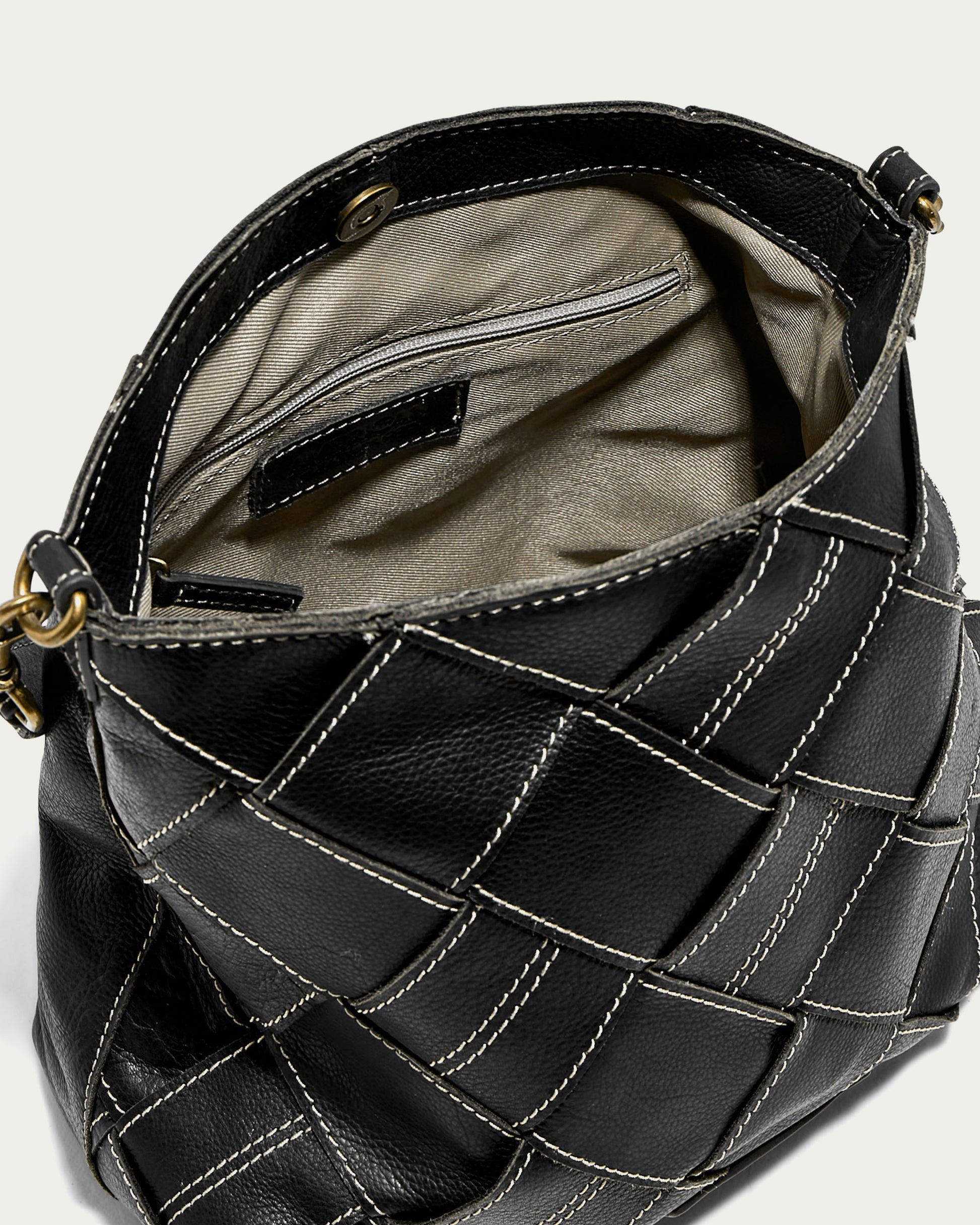 Close-up of an open black Mercer Shoulder handbag by American Leather Co., showcasing a patchwork design with white stitching. Made from genuine leather, it features a beige interior with a small zippered pocket. The bag is equipped with a snap closure at the top and includes a detachable shoulder strap for versatile wear.