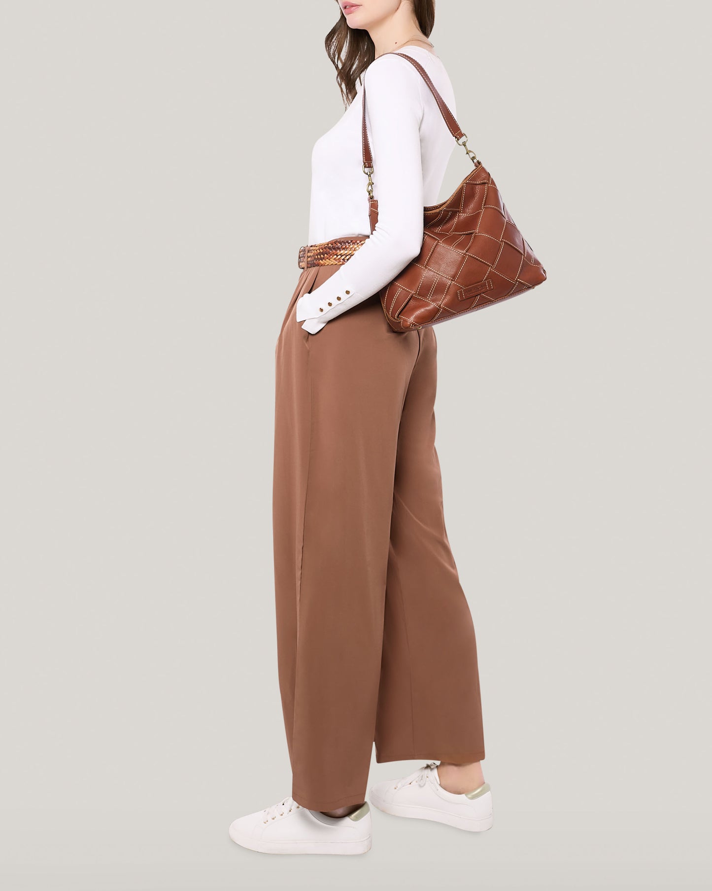 A person is dressed in a white long-sleeve top with button details on the sleeves, brown high-waisted wide-leg pants, and white sneakers. They are carrying the Mercer Shoulder handbag by American Leather Co., a large genuine leather bag with quilted stitching and a detachable shoulder strap, over their shoulder, standing against a plain light background.