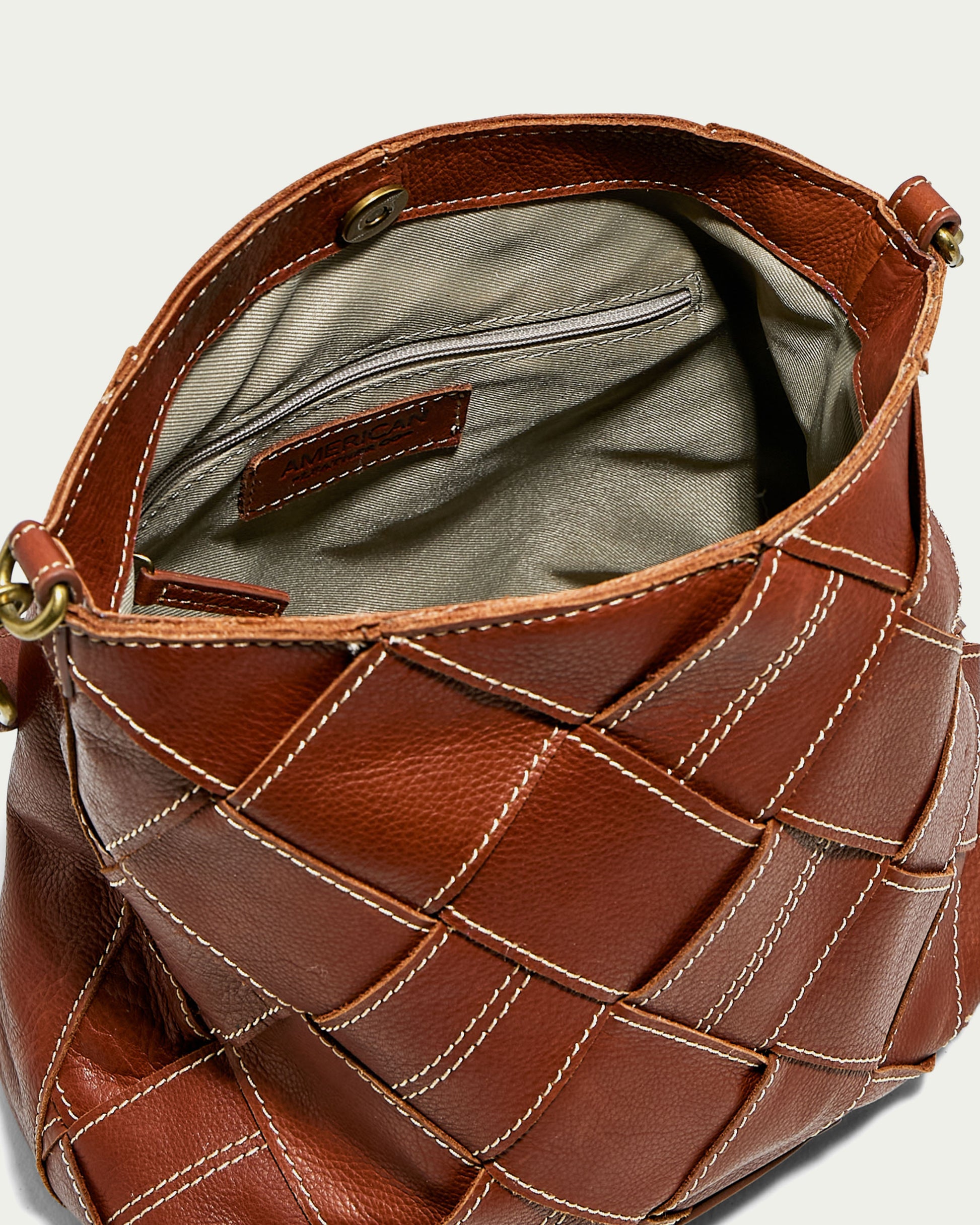 The Mercer Shoulder handbag by American Leather Co. showcases its open interior, revealing a beige lining and an inner zipped pocket. Crafted from genuine brown leather with a woven pattern and accented by white stitching, the bag features metal rings on the sides to attach the detachable shoulder strap.