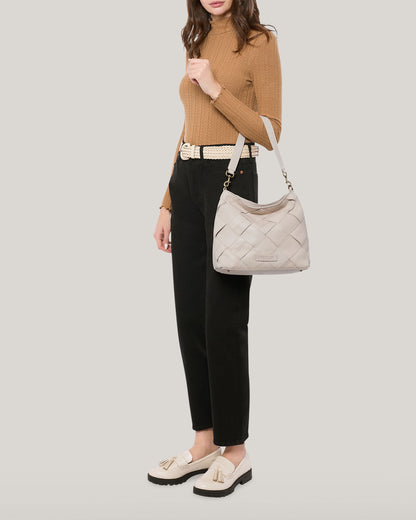 A person dressed in a brown turtleneck and black pants holds the stylish Mercer Shoulder bag by American Leather Co., featuring a detachable strap. Their look is completed with white loafers, exuding effortless chic against the plain light grey background.