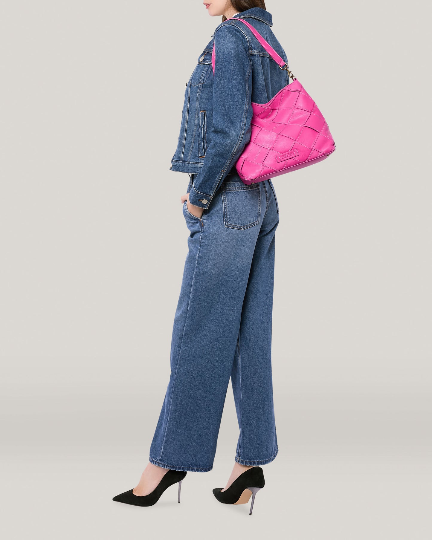 A person dressed in a blue denim jacket and jeans, paired with black high-heeled shoes, stands holding the bright pink Mercer Shoulder handbag from American Leather Co., featuring a genuine leather detachable shoulder strap, over their shoulder. The background is plain and light-colored. The pose is casual and stylish.