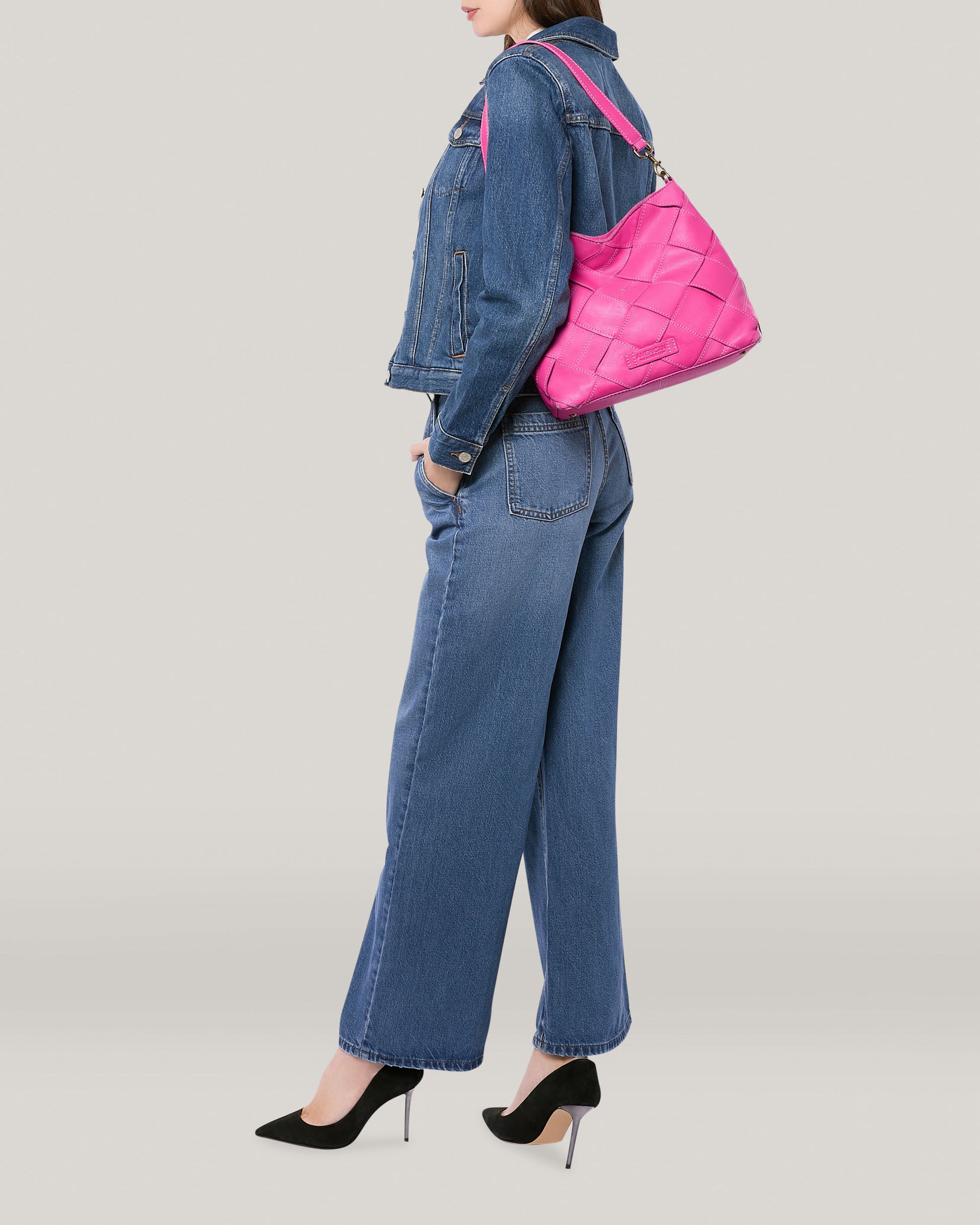 A person dressed in a blue denim jacket and jeans, paired with black high-heeled shoes, stands holding the bright pink Mercer Shoulder handbag from American Leather Co., featuring a genuine leather detachable shoulder strap, over their shoulder. The background is plain and light-colored. The pose is casual and stylish.