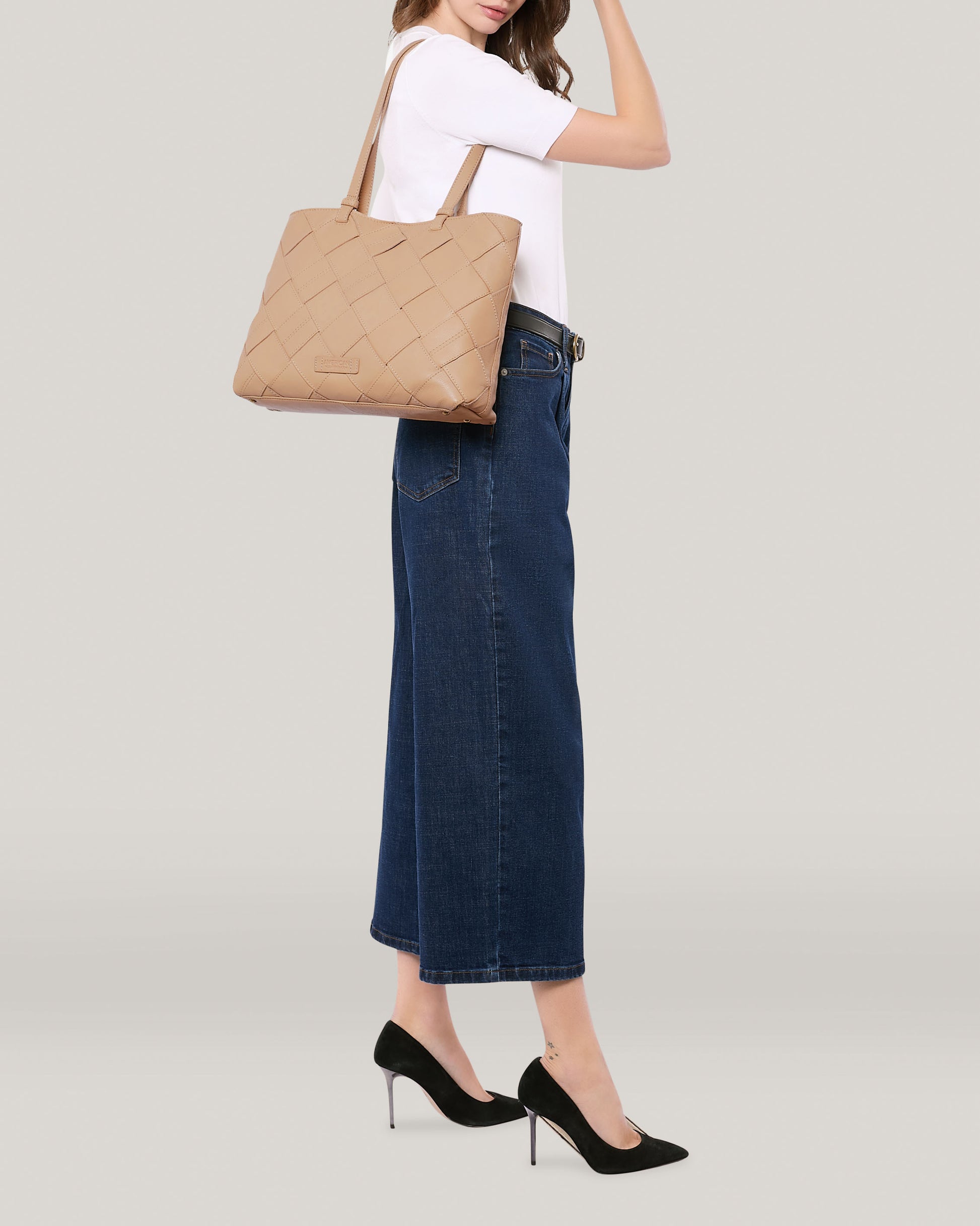 A person dressed in a white t-shirt, long blue jeans, and black heels carries the Mercer Woven Tote by American Leather Co., featuring hand-weaved panels in a large tan design, slung over their shoulder against a plain, light-colored background.