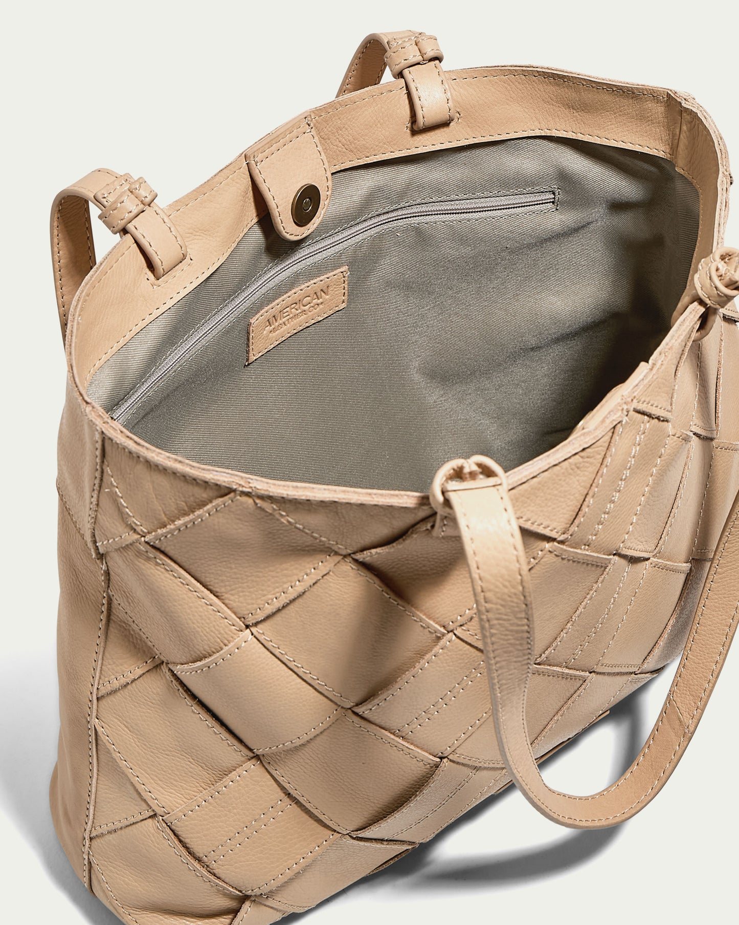 The Mercer Woven Tote by American Leather Co. is displayed open, displaying its straightforward interior. The bag is crafted from genuine leather in a beige color, showcasing a quilted pattern. It includes dual handles and a snap closure, with the brand label visible inside.