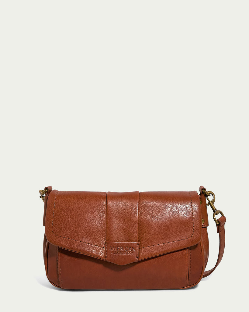 The Mohegan Crossbody is a brown leather shoulder bag designed with a magnetic flap closure and an adjustable strap, crafted from genuine leather. The brand name "American Leather Co." is embossed on a tag on the front.