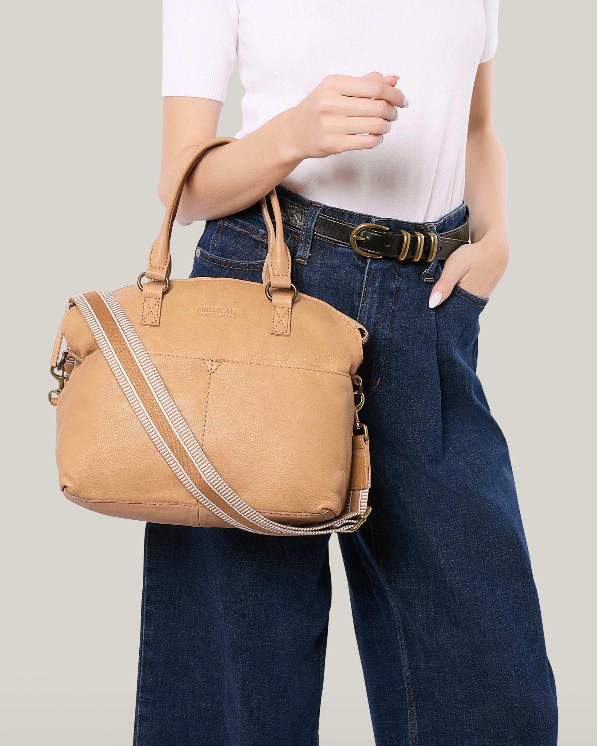 A person in a white top and high-waisted blue jeans carries a tan leather handbag from American Leather Co., featuring a striped design and antiqued hardware. The bag has both handles and a detachable Striped Web Strap, while the person casually slips a hand into their pocket.