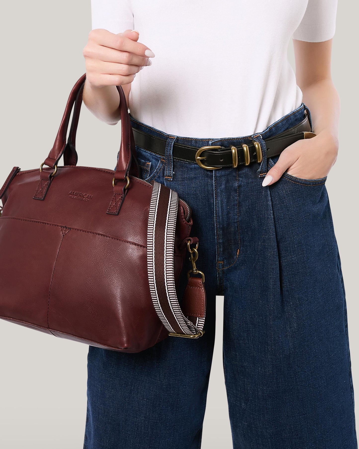 An individual dressed in a white shirt and blue jeans is carrying an American Leather Co. handbag, which has a brown leather finish and features an adjustable Striped Web Strap. The bag is adorned with elegant antiqued hardware. They are also wearing a black belt accented by a stylish gold buckle, with their hand resting casually in a pocket.