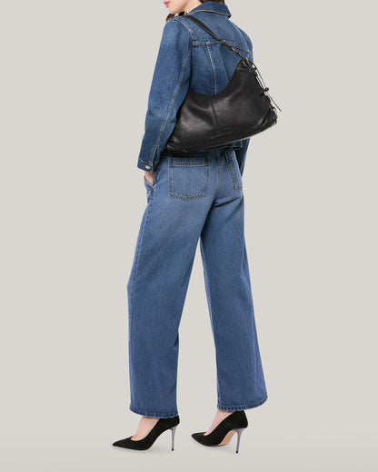 A person stands against a plain background, facing slightly away from the camera. They are wearing a blue denim jacket and matching wide-leg jeans, paired with black stiletto heels. Over their shoulder is an American Leather Co. Thayer Perfect Hobo leather purse.