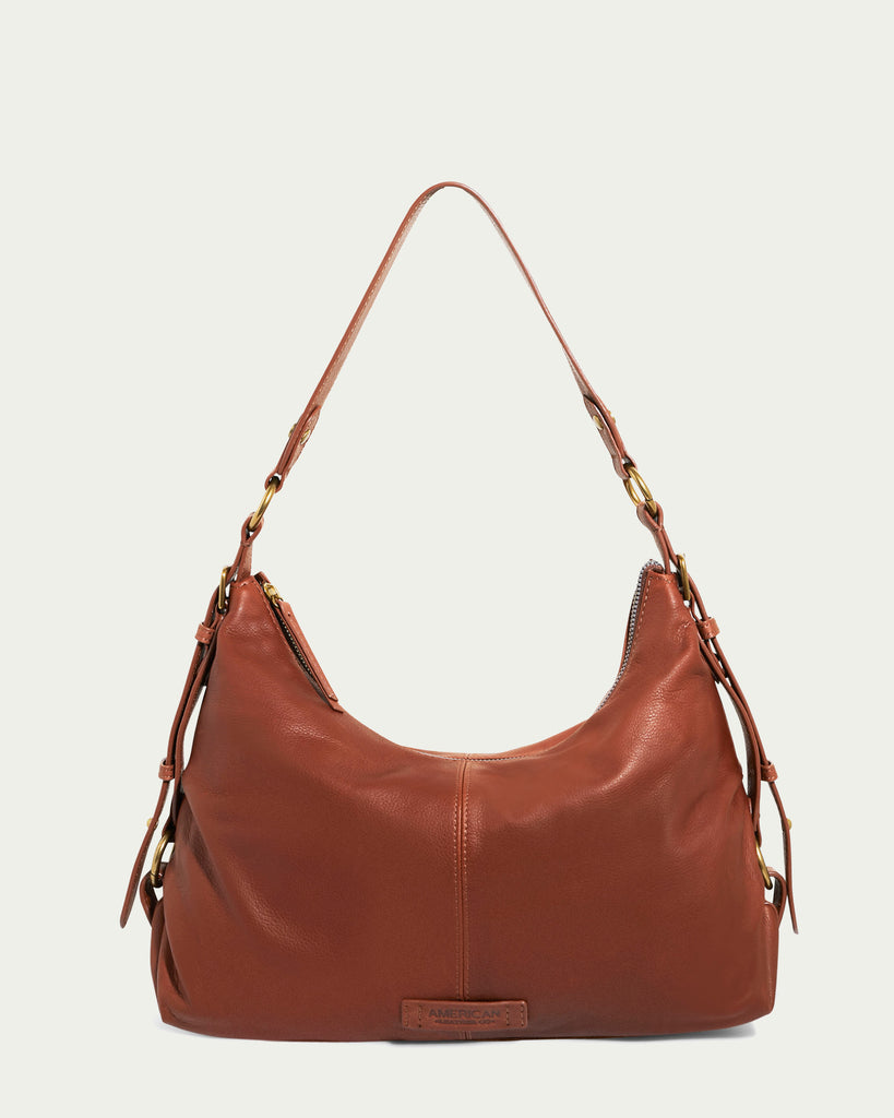 Thayer Perfect Hobo by American Leather Co.: A brown genuine leather hobo handbag featuring a single shoulder strap and gold hardware. The bag includes side buckles and a visible brand label near the bottom. Its sleek and minimalist design boasts a curved top and a spacious main compartment.