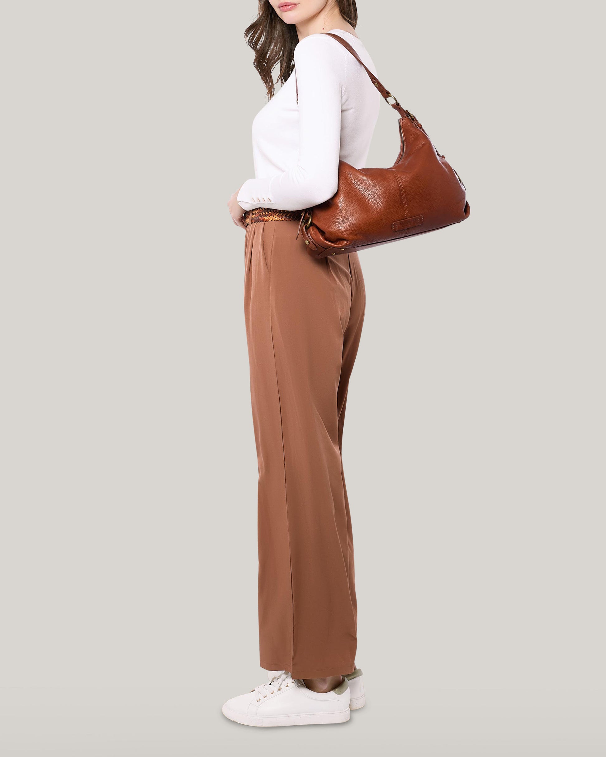 A woman stands sideways against a plain, light gray background. She is wearing a white long-sleeve top, high-waisted brown trousers, and white sneakers. She has a medium-sized Thayer Perfect Hobo leather purse from American Leather Co. slung over her shoulder.