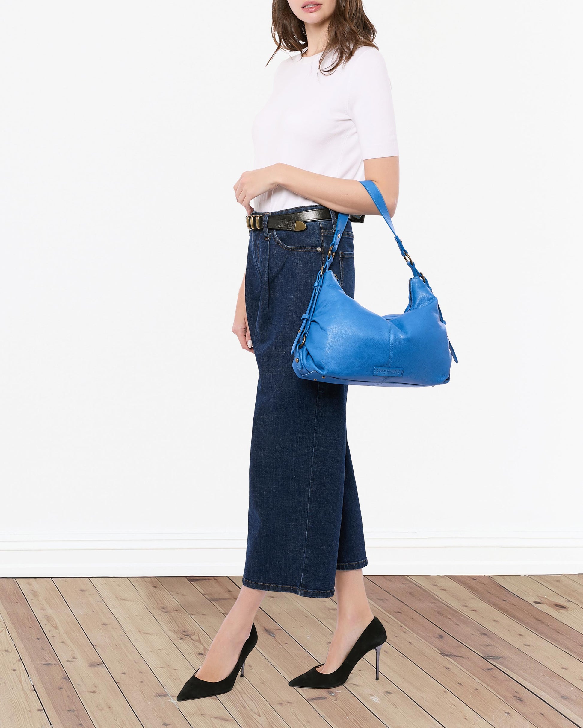 A woman on a wooden floor wears a white shirt, blue jeans, and black heels. Draped over her shoulder is the American Leather Co.'s Thayer Perfect Hobo, a bright blue genuine leather handbag. Her face remains hidden in the image.