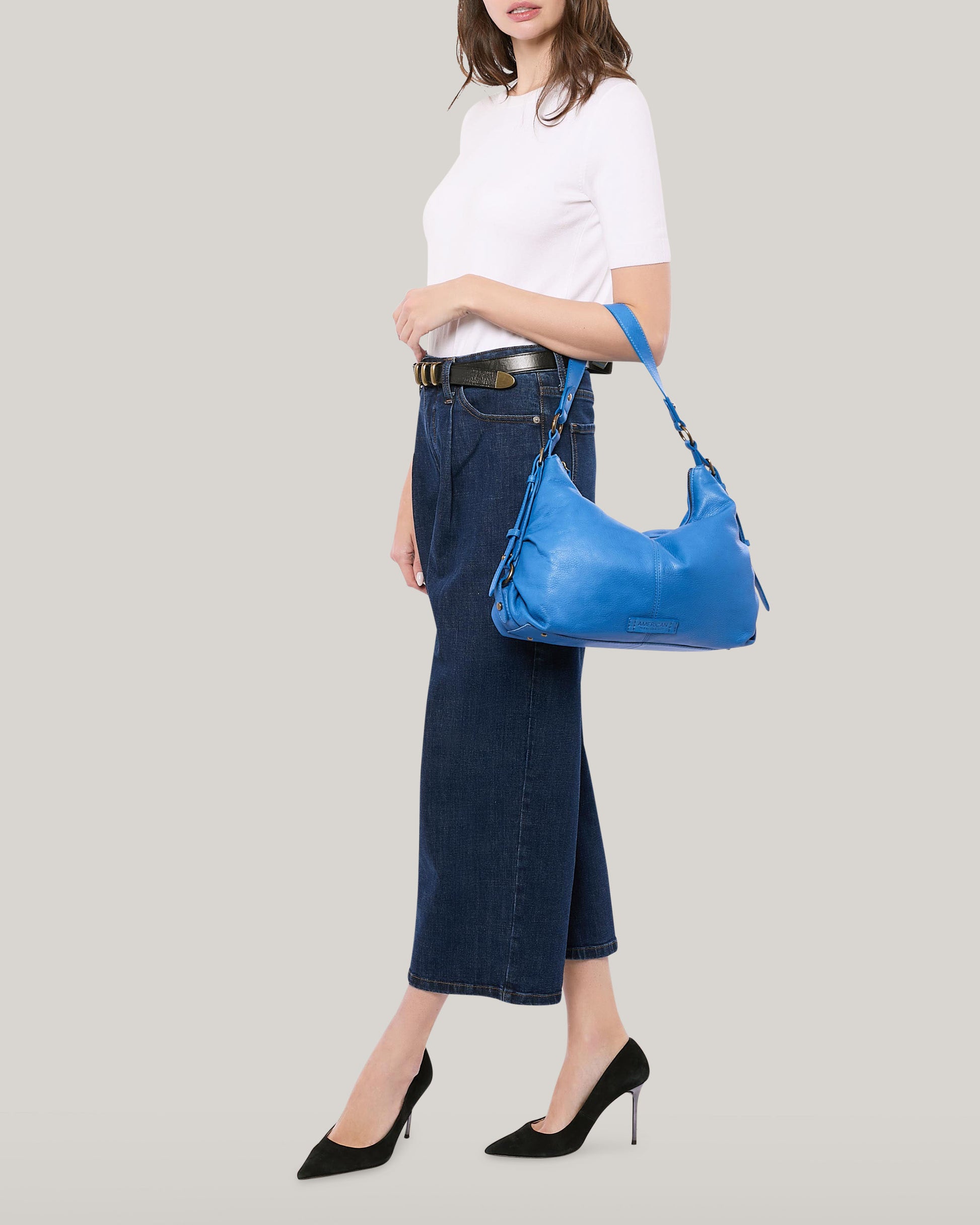 A person dressed in a white t-shirt, dark denim jeans, and black heels carries the Thayer Perfect Hobo by American Leather Co., known for its bright blue genuine leather. The backdrop is a plain light gray.