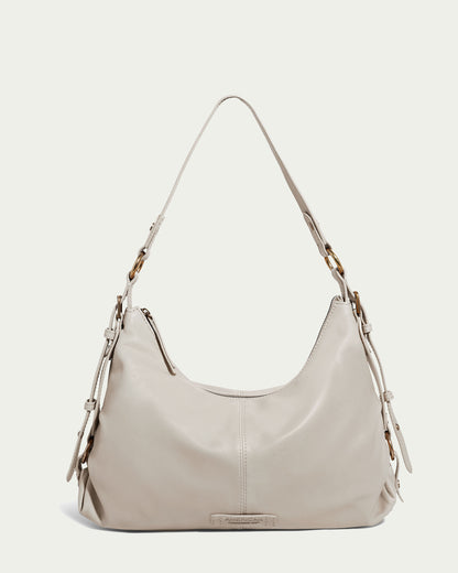 Discover the Thayer Perfect Hobo by American Leather Co.—a cream-colored leather purse with a distinctive crescent shape. It features a shoulder strap accented with brown metal buckles. This genuine leather handbag showcases an elegant minimalist design, a smooth texture, and includes a small rectangular tag at the bottom center.