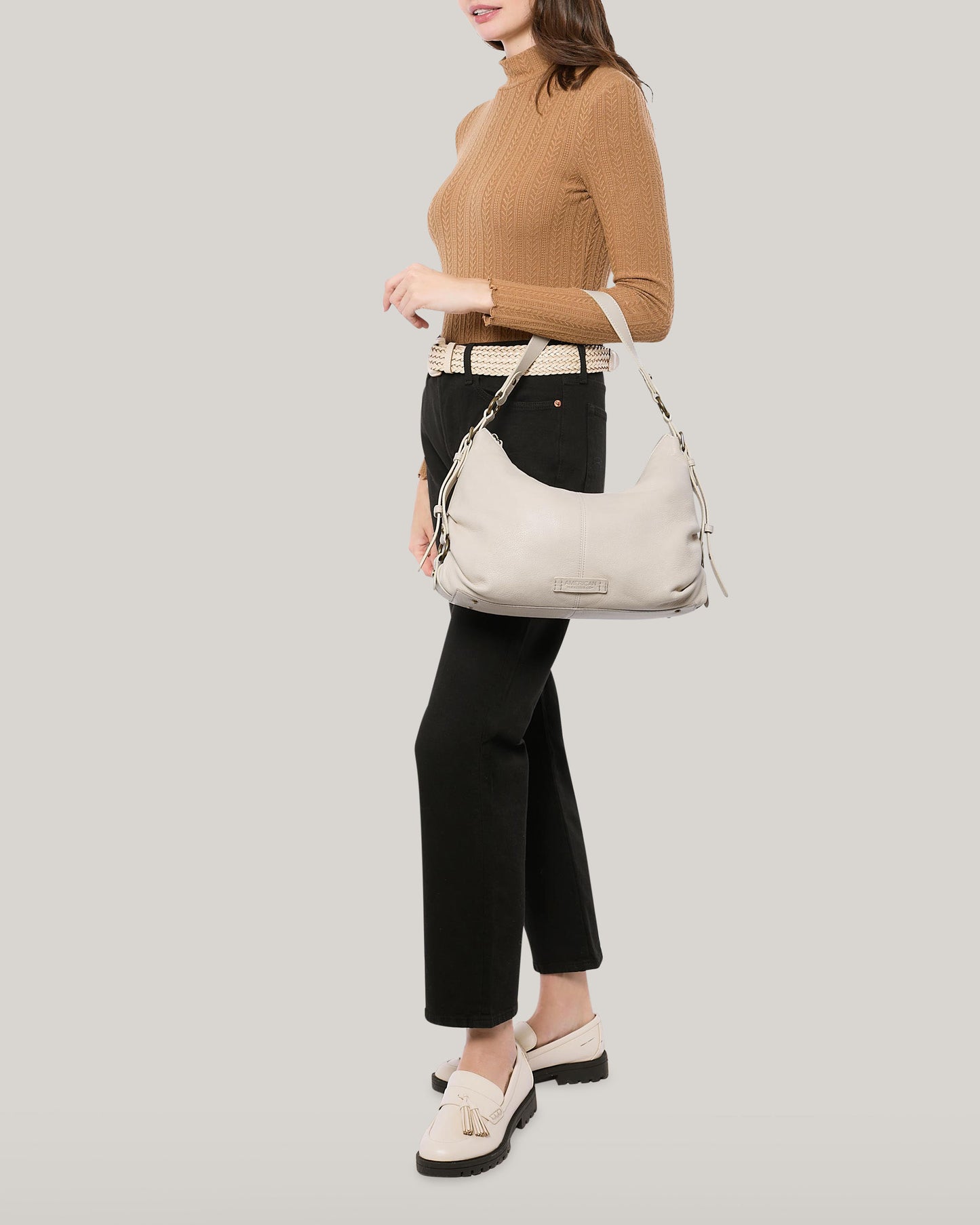 A woman in a brown sweater and black pants holds an American Leather Co. Thayer Perfect Hobo, a cream-colored leather purse. She has on beige shoes with tassels and a matching belt, set against a plain light gray background.