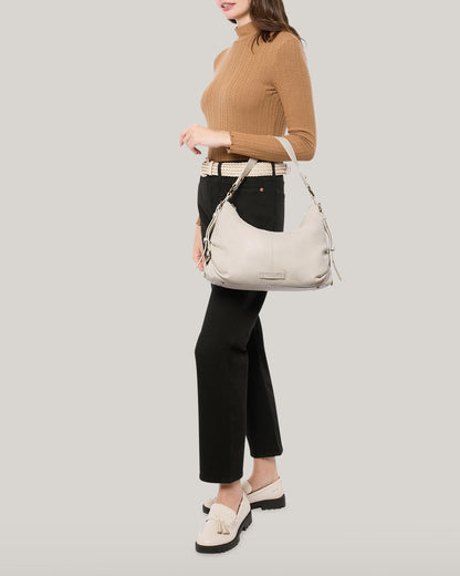 A woman in a brown sweater and black pants holds an American Leather Co. Thayer Perfect Hobo, a cream-colored leather purse. She has on beige shoes with tassels and a matching belt, set against a plain light gray background.