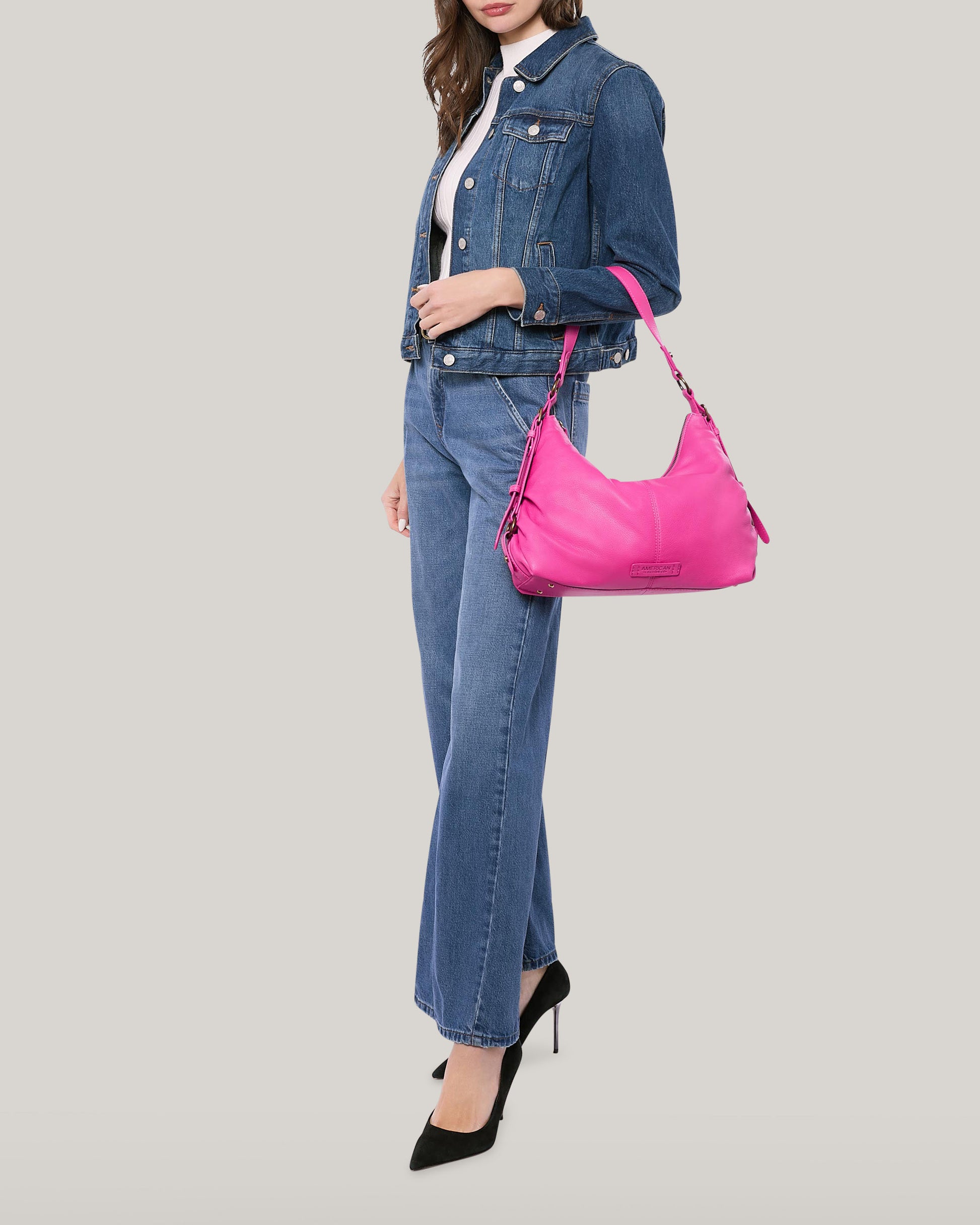 A person wearing a denim jacket and jeans, a white top, and black high heels is holding a bright pink American Leather Co. Thayer Perfect Hobo on their left shoulder. The background is a plain, light neutral color. The genuine leather handbag stands out due to its vibrant color.