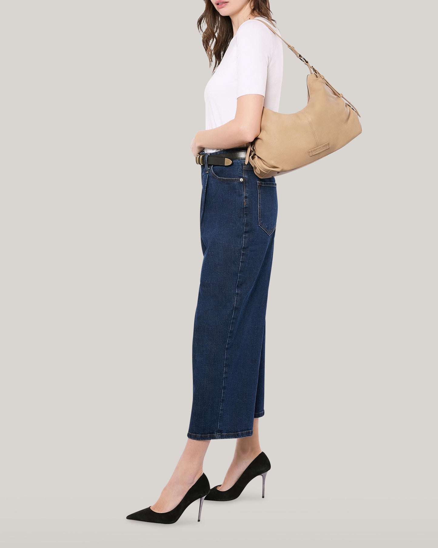 A person dressed in a white t-shirt, blue denim skirt, and black high heels is holding the Thayer Perfect Hobo from American Leather Co., a tan shoulder bag made of genuine leather. The background is plain light gray.