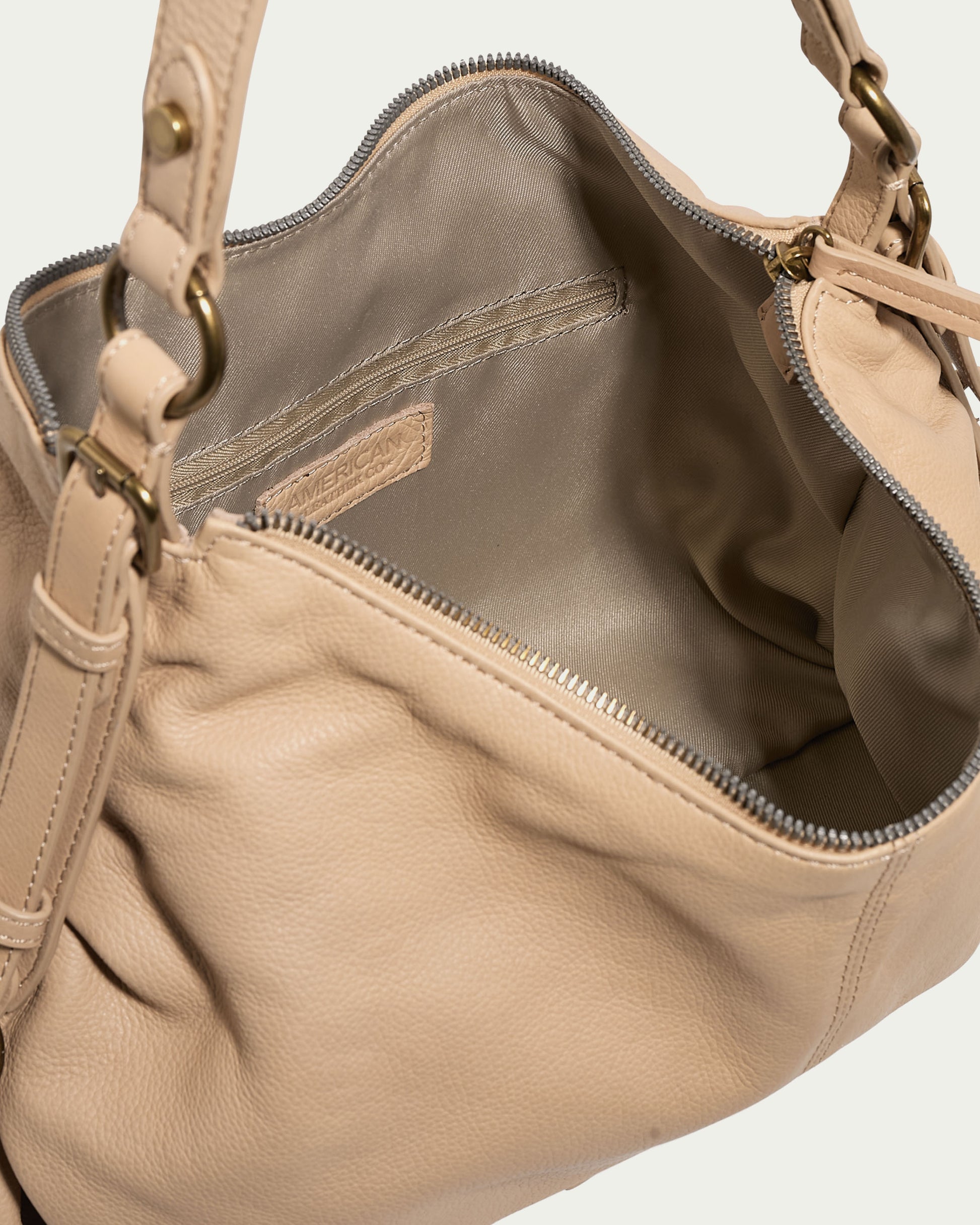 The Thayer Perfect Hobo by American Leather Co. is an open beige leather handbag featuring a visible interior lining and a metal zipper. It showcases an eye-catching inside pocket, complemented by its strong handles with metal attachments, embodying the essence of genuine leather craftsmanship.