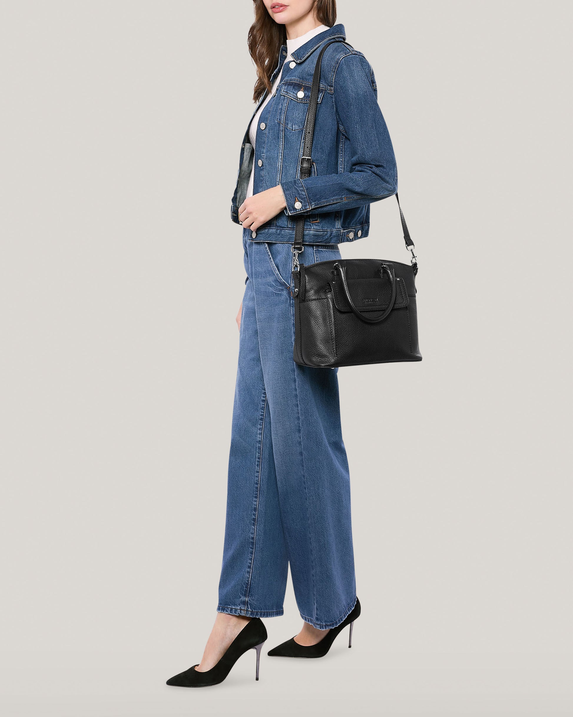 A person stands posing against a light grey background. They are wearing a denim jacket, a white top, wide-leg denim jeans, and black high heels. Over their shoulder, they carry the chic Vona Convertible Dome Satchel in genuine leather by American Leather Co.