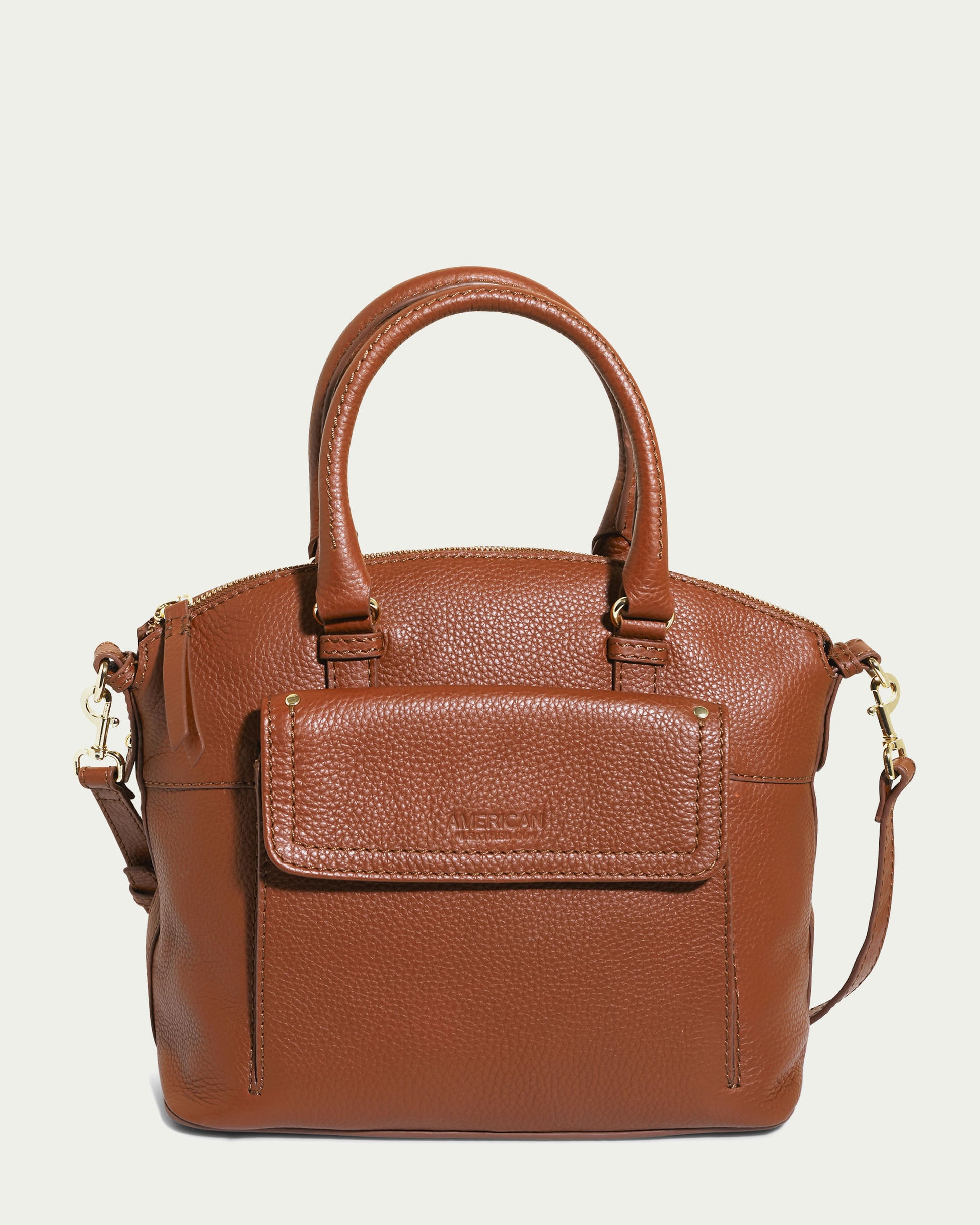 A Brandy Pebble Vona Convertible Dome Satchel from American Leather Co., featuring two short, rounded handles and a detachable shoulder strap. This genuine leather handbag includes a front flap pocket with a magnetic closure and gold-tone hardware, such as zippers and strap clasps. The bag boasts a structured, textured design.