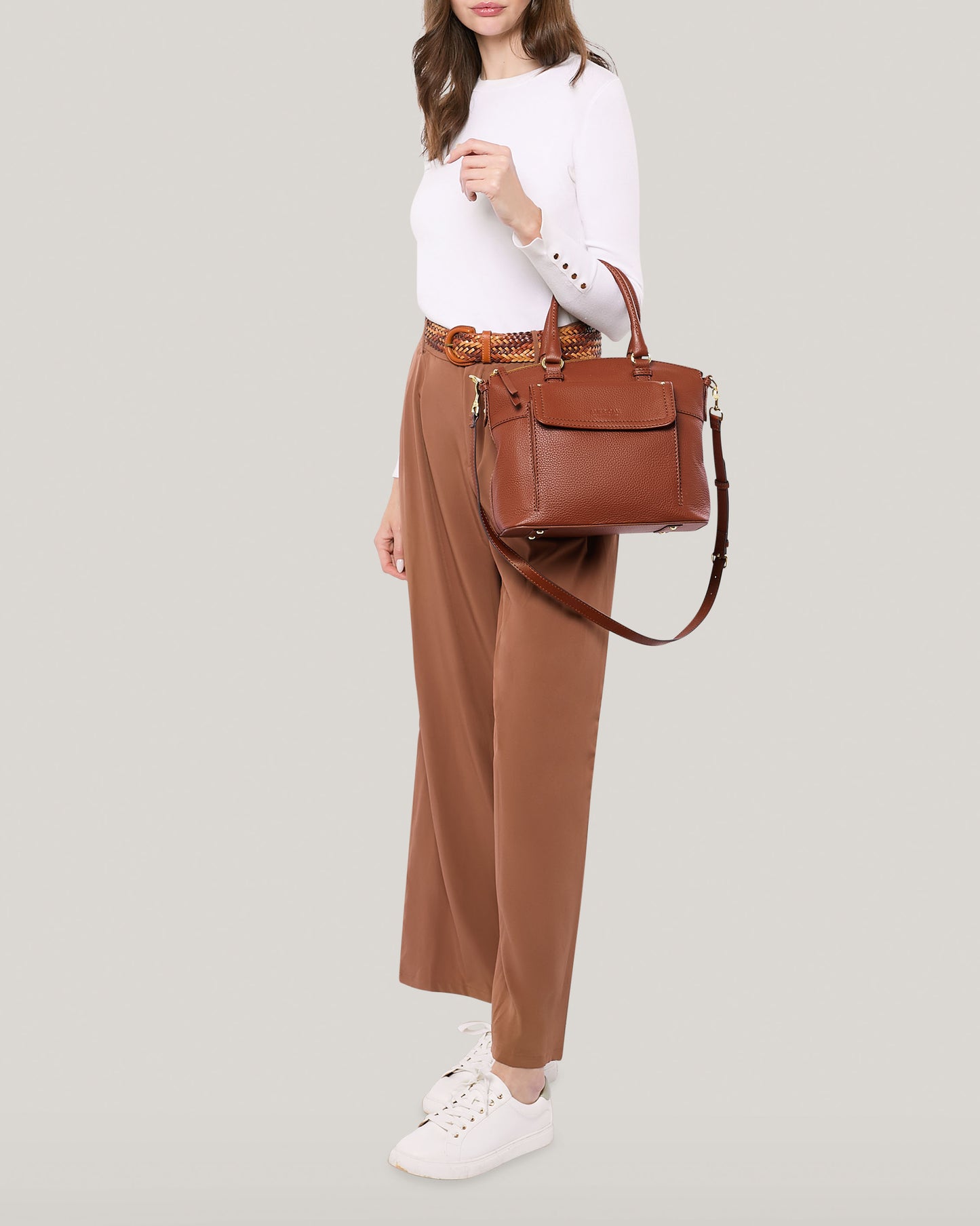 A woman wearing a white long-sleeve blouse and tan high-waisted trousers with a braided brown belt stands against a light background. She carries the Vona Convertible Dome Satchel by American Leather Co., made of genuine leather with a shoulder strap, and wears white sneakers.