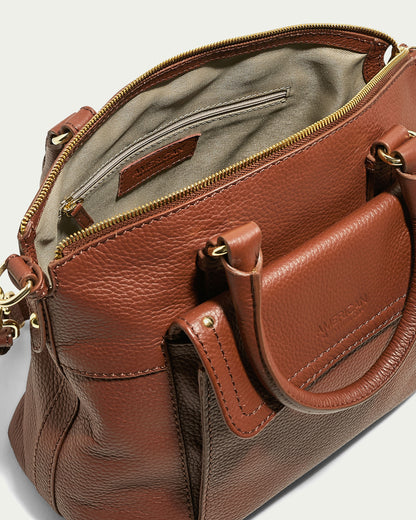 The Vona Convertible Dome Satchel by American Leather Co. is a brown Forever Pebble leather handbag with a pebble texture, gold-toned hardware, dual handles, and a detachable shoulder strap. This versatile satchel opens to reveal a beige fabric interior, featuring a zipper pocket and an interior brand label.