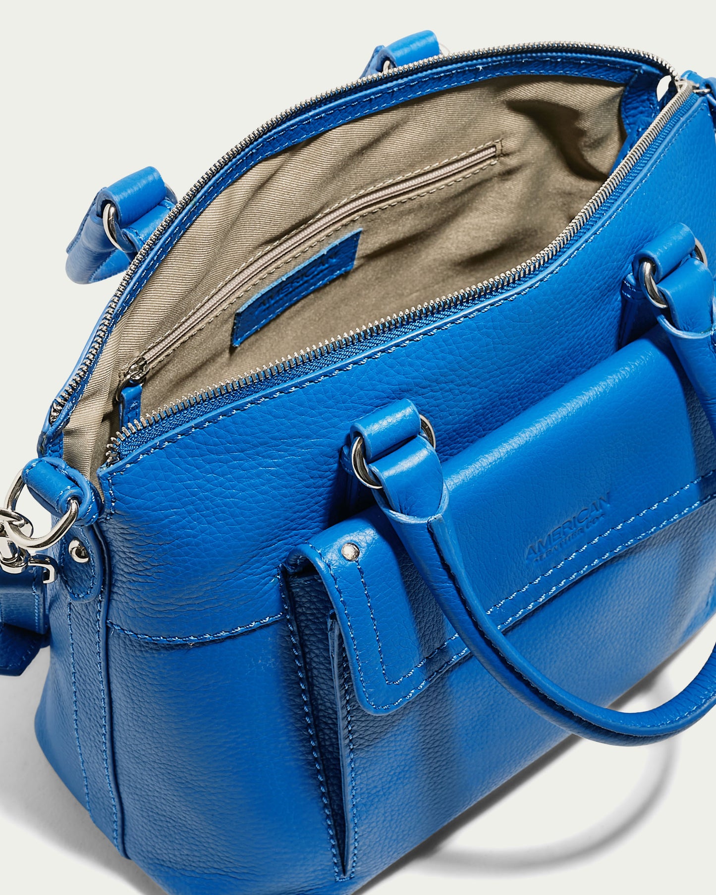 A vibrant blue Vona Convertible Dome Satchel by American Leather Co. with silver hardware opens to reveal its beige interior. The genuine leather bag features two handles, a detachable shoulder strap, a front pocket with a flap, and an interior zippered pocket. The brand name is embossed on the front.