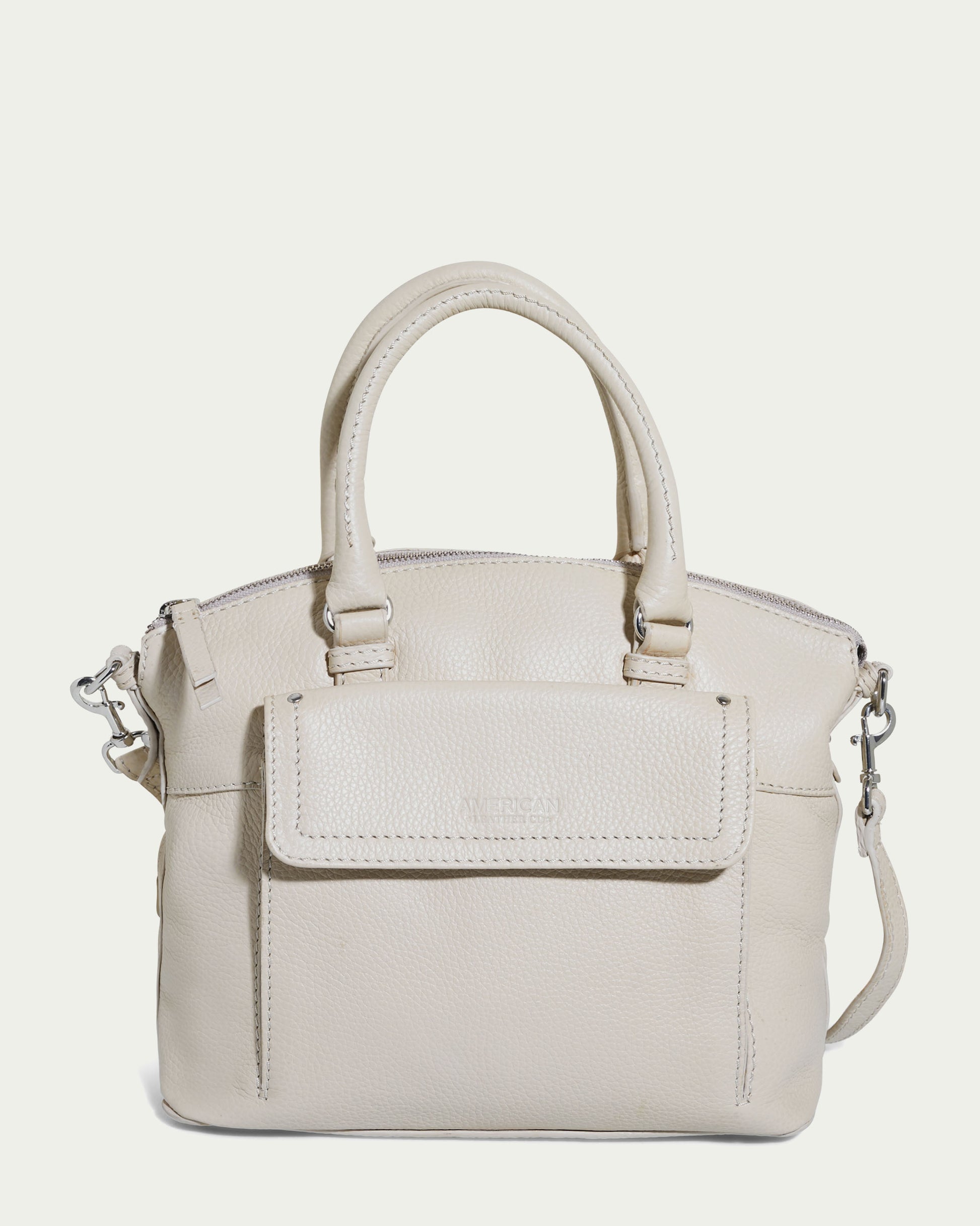 The Vona Convertible Dome Satchel by American Leather Co. comes in a light beige "Ecru Pebble" genuine leather with dual handles and an adjustable, detachable shoulder strap. This handbag features a front flap pocket, zipper closure, and silver hardware, with a smooth texture and slightly pebbled finish.