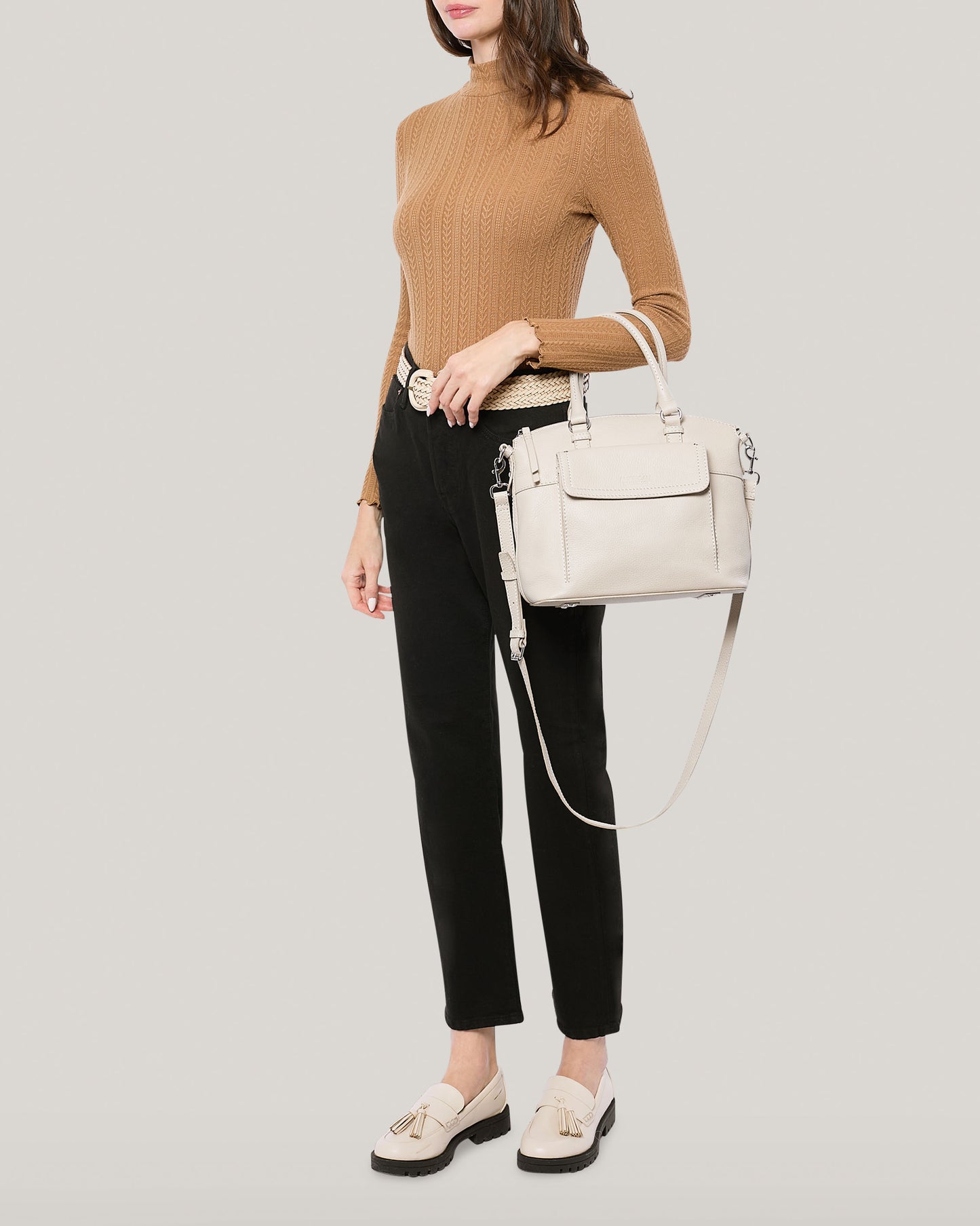 A person dressed in a long-sleeve camel-colored sweater, black pants, white belt, and white loafers with tassels is holding the Vona Convertible Dome Satchel by American Leather Co., which features an adjustable strap. The background is plain light grey.