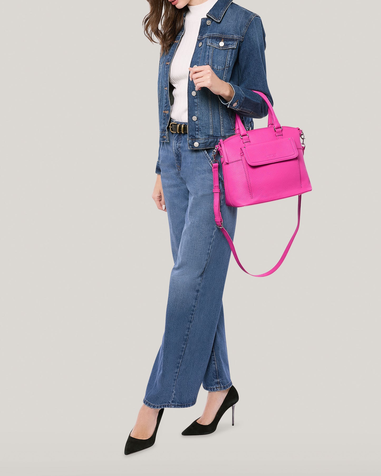 A woman dressed in a denim jacket and jeans, white turtleneck, and black heels holds a bright pink Vona Convertible Dome Satchel from American Leather Co. She stands against a plain light grey background. The genuine leather handbag features both a short handle and a long strap. Her face is not visible in the image.
